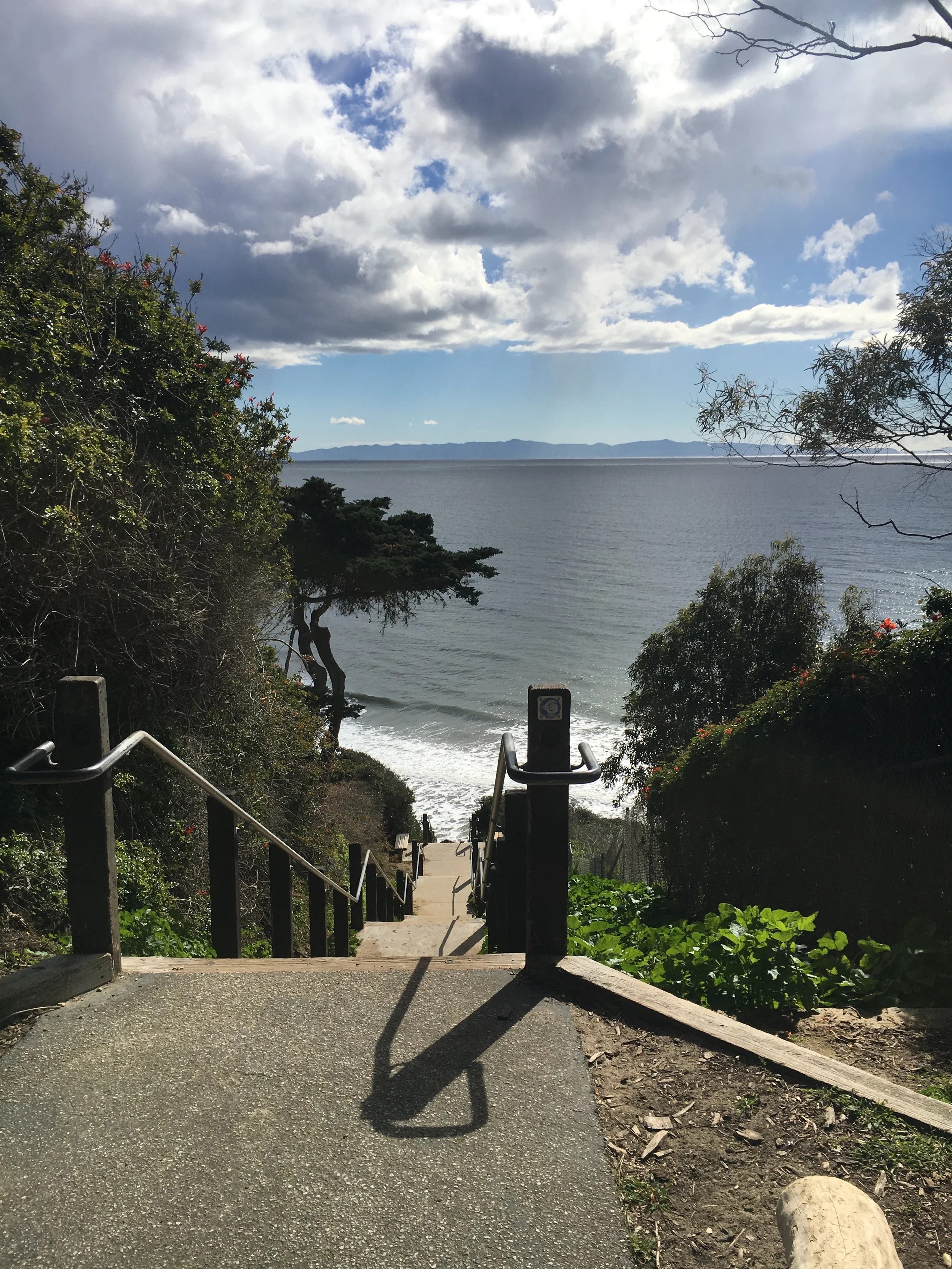 A marketing photo of a Santa Barbara staircase leading down towards Hendry's Beach on the Pacific Ocean, with trees on either side and a cloudy sky overhead the Channel Islands.