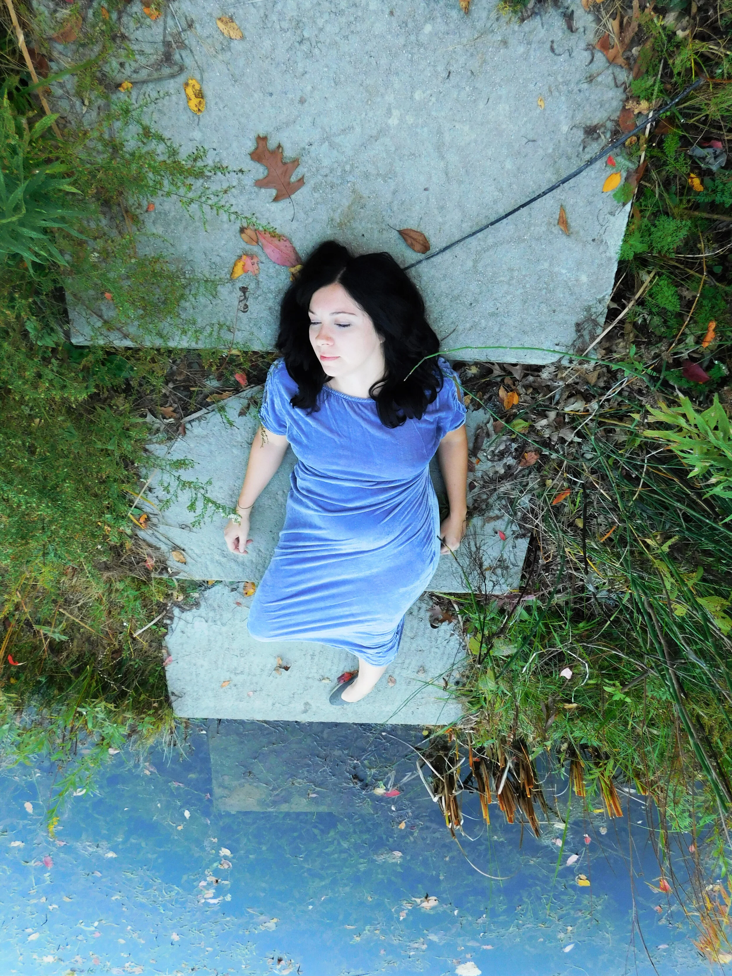 A person in a long velvet purple dress and black hair reclines on cement, leaf-strewn steps, half in a lake of water.