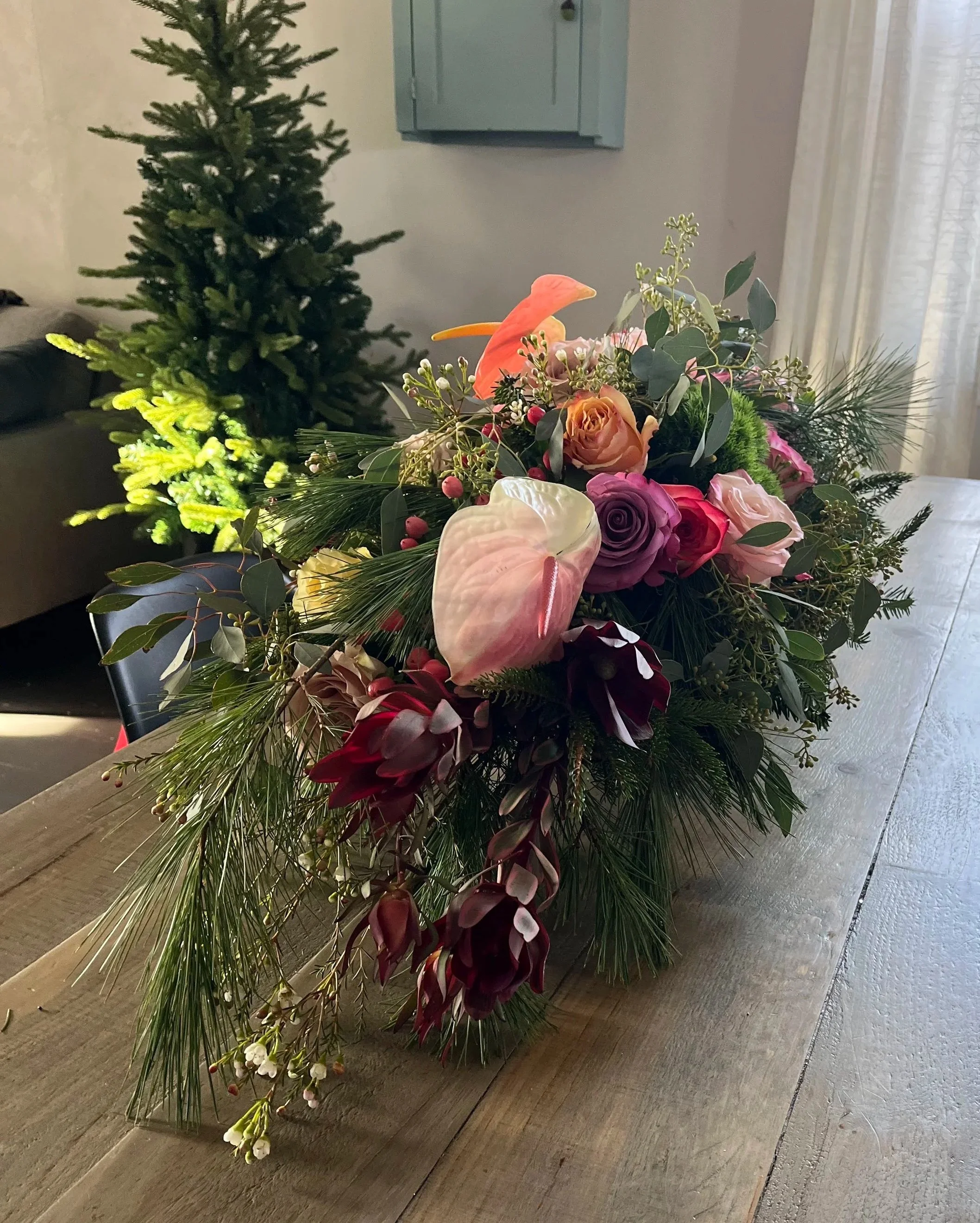 Close up of a colorful, long, low floral arrangement with roses, anthuriums, waxflower, hypericum and holiday greenery.