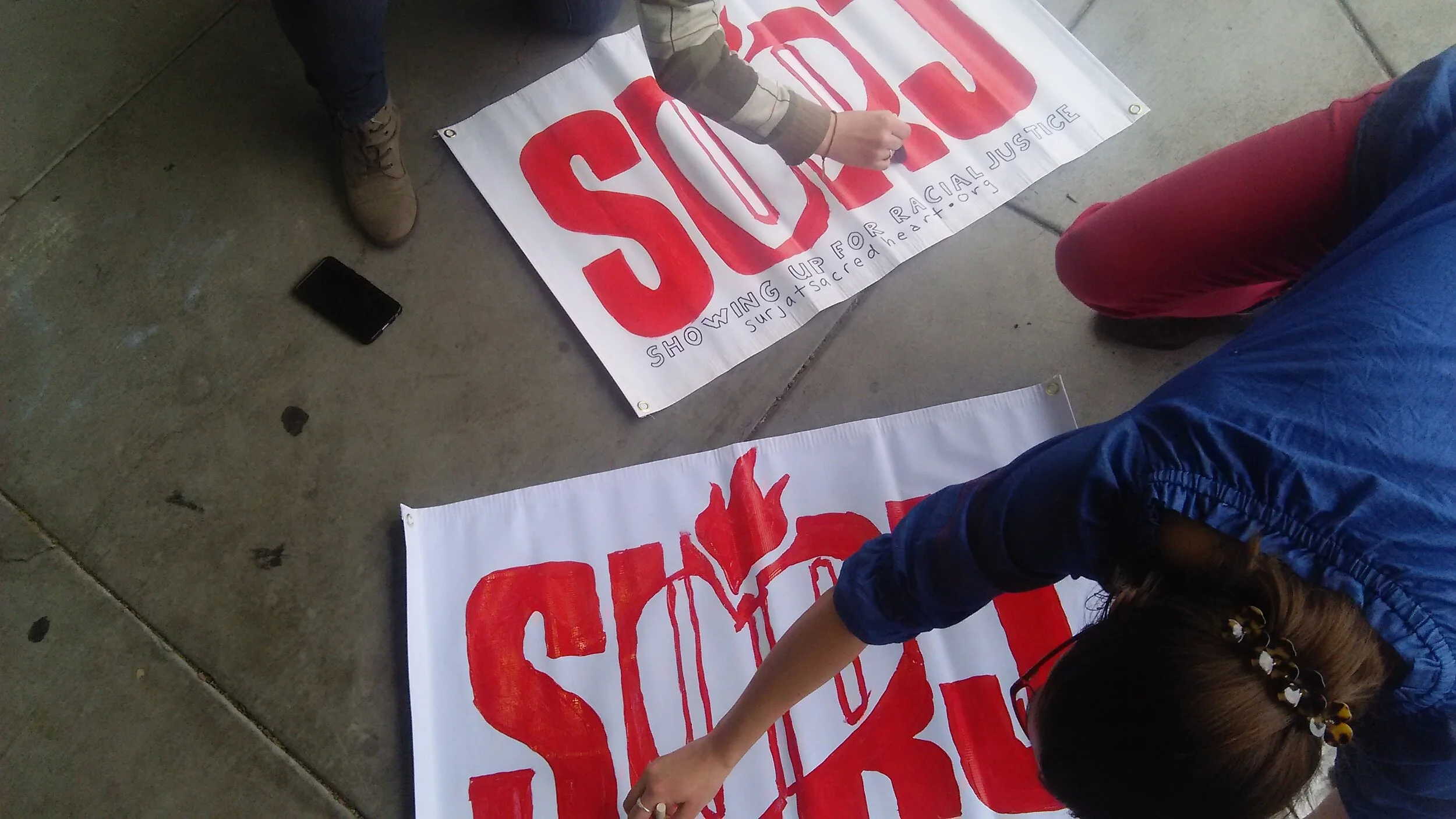 Photograph taken from above showing two people crouched over white banners onto which they are hand painting the SURJ logo in red