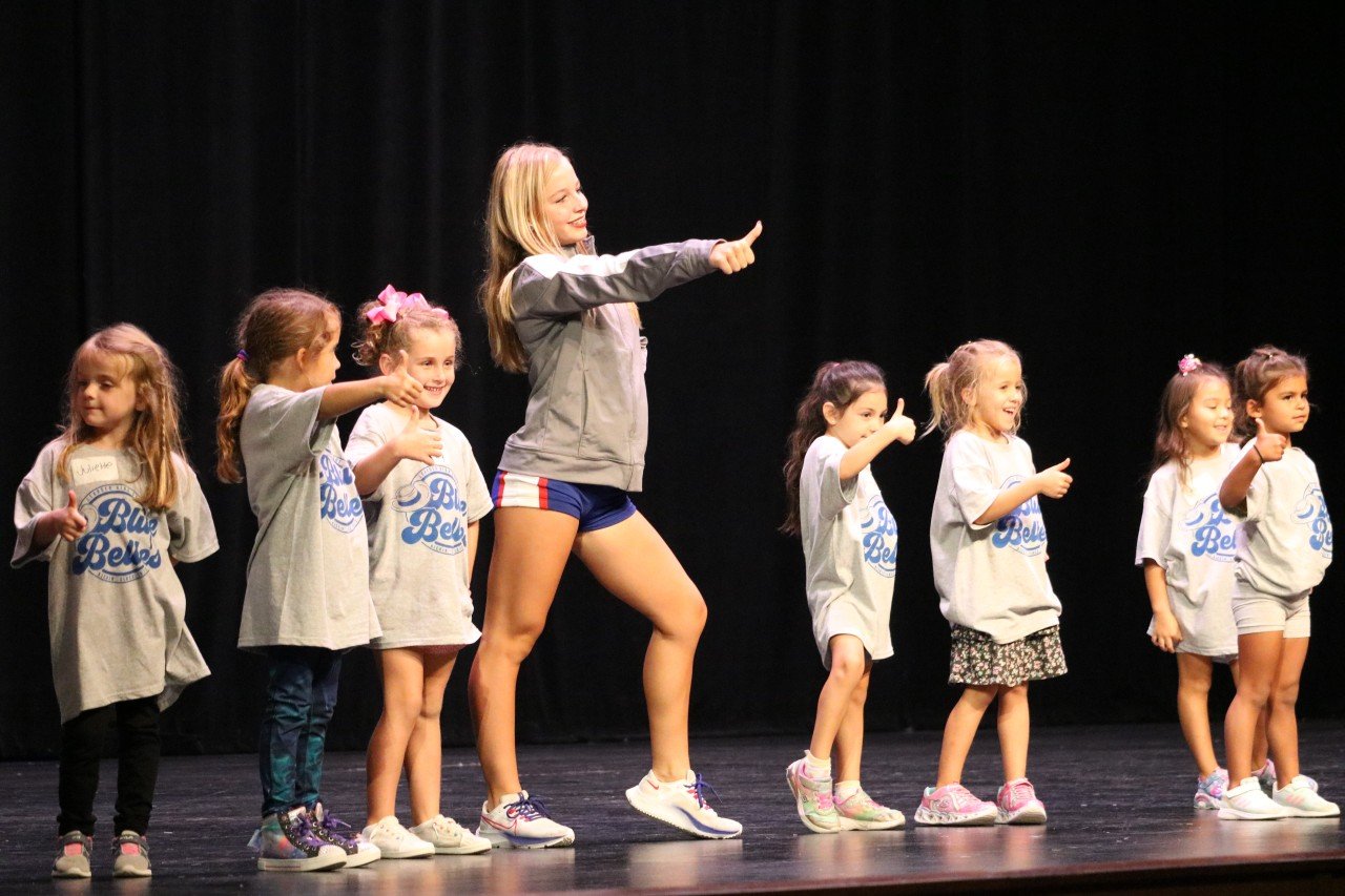 A group of young children and a teenager on stage participating in a dance or cheerleading event, wearing matching gray shirts with a "Blue Belles" logo. The children are mimicking the teenager's pose, all giving a thumbs-up gesture.