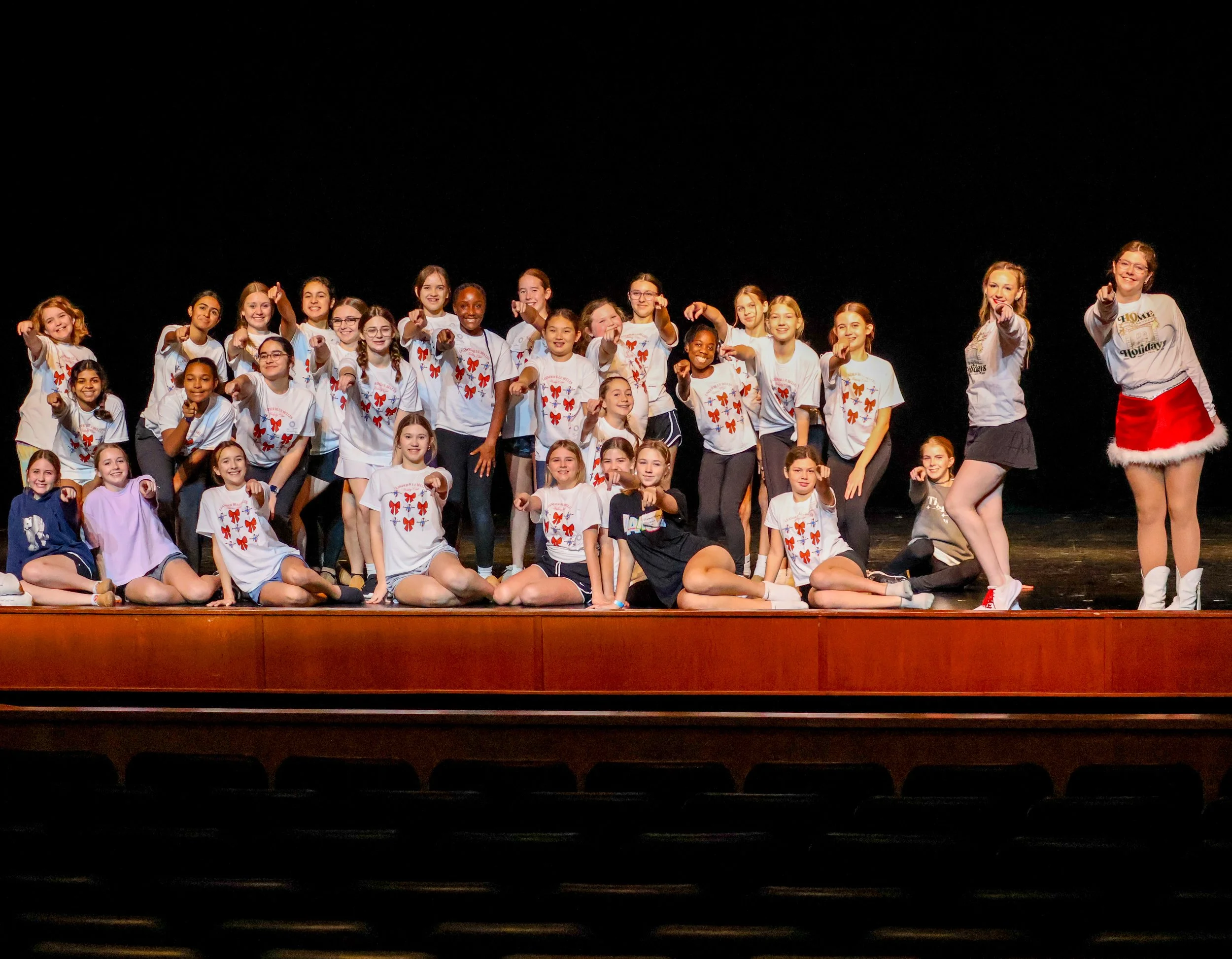 Group of Dance Clinic participants on stage, posing for a photo, with a black background and a wooden stage. Performing at the LHS Blue Belles Holiday Show