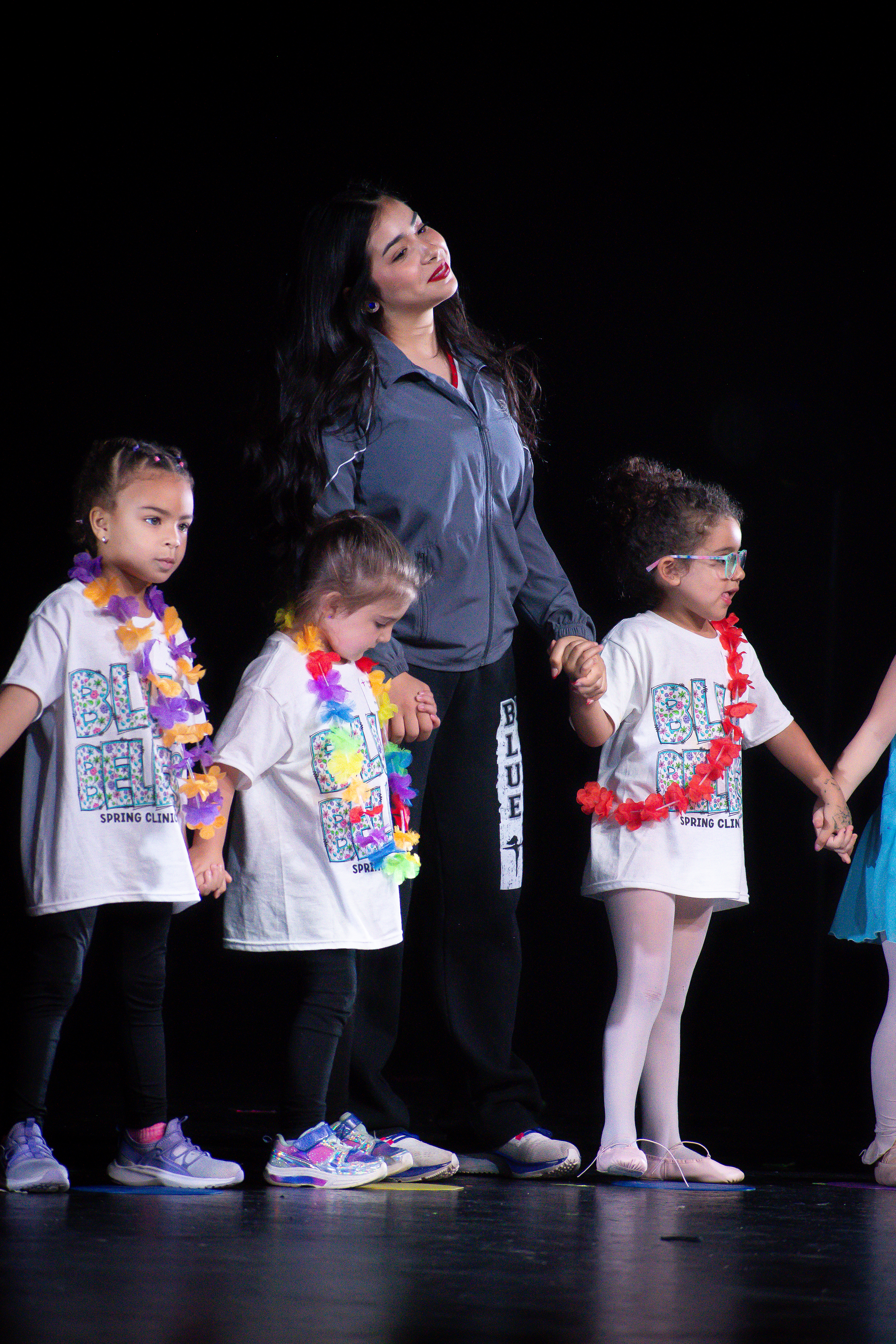 A Blue Belle and four young girls holding hands on stage, participating in a Dance Clinic. The girls are wearing white shirts with colorful designs, leis, and leggings or tights, with the woman dressed in a gray jacket and black pants. Drill team