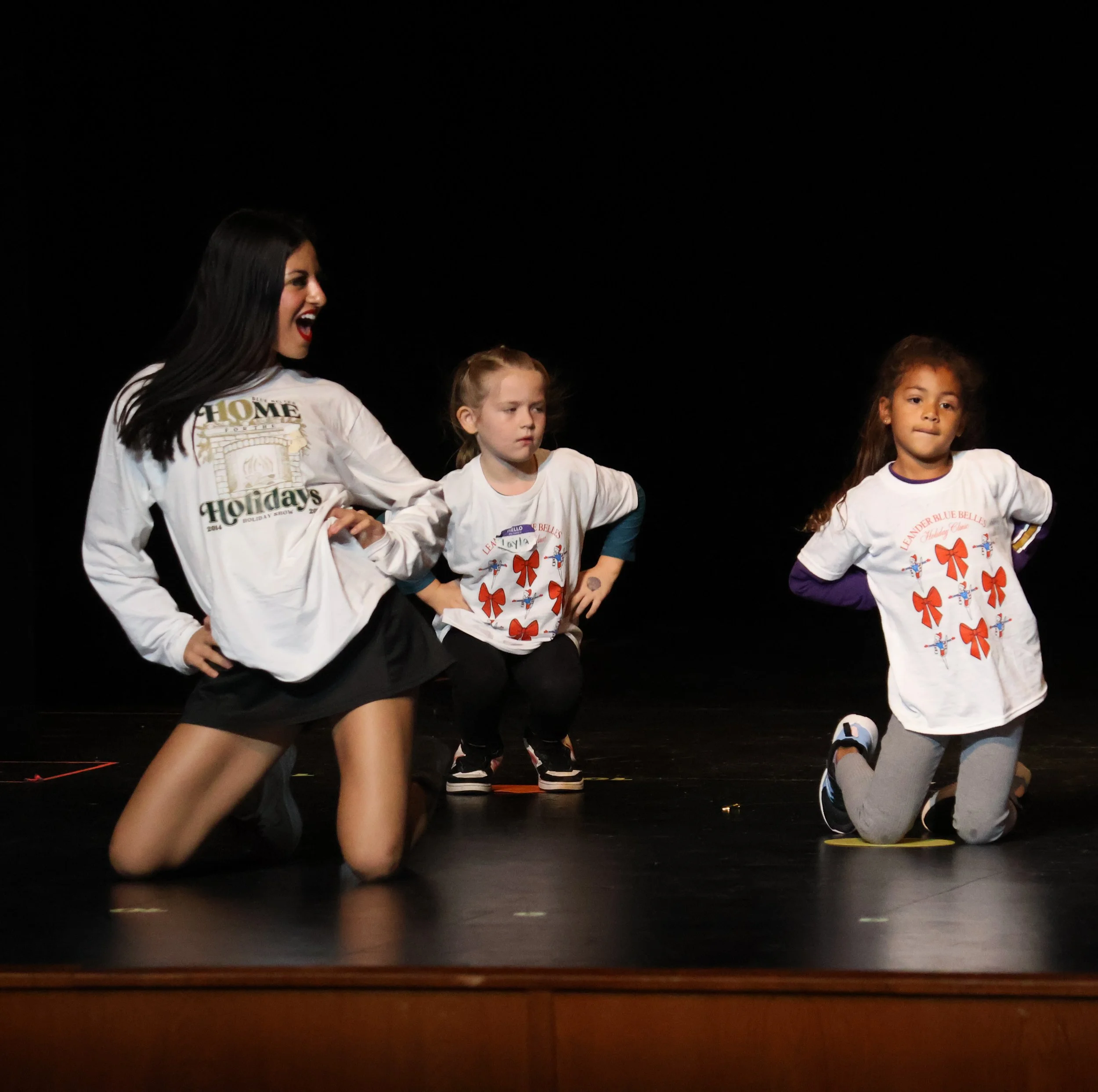 Three young girls and a woman performing on stage during a dance event, all wearing white T-shirts with festive red bows. Smiling and posing with her hands on her hips, while the girls are kneeling, inside the LHS PAC at a Blue Belles Dance Clinic