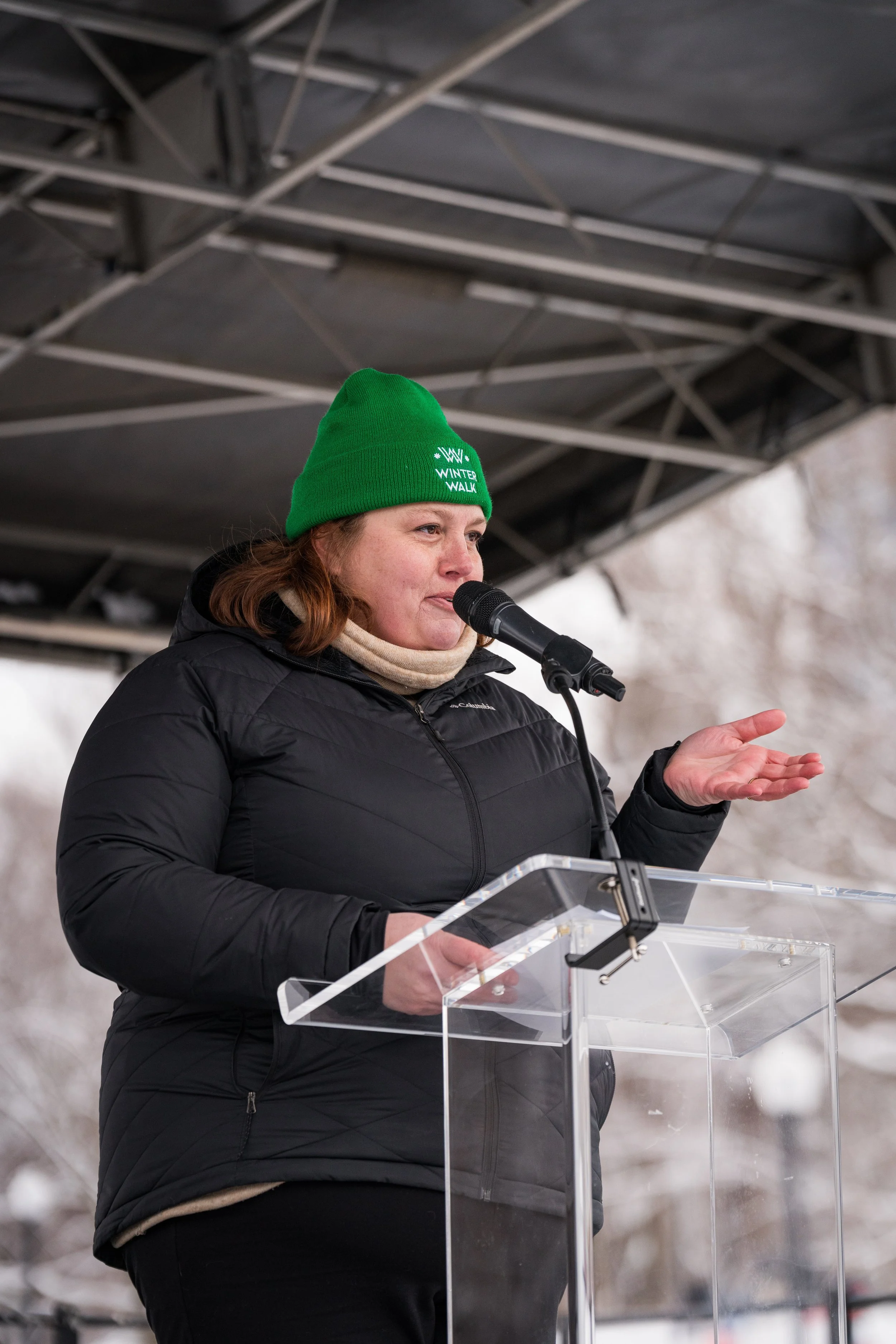 Meag is wearing a winter walk beanie and her puffer jacket, on stage with a microphone at a podium while speaking