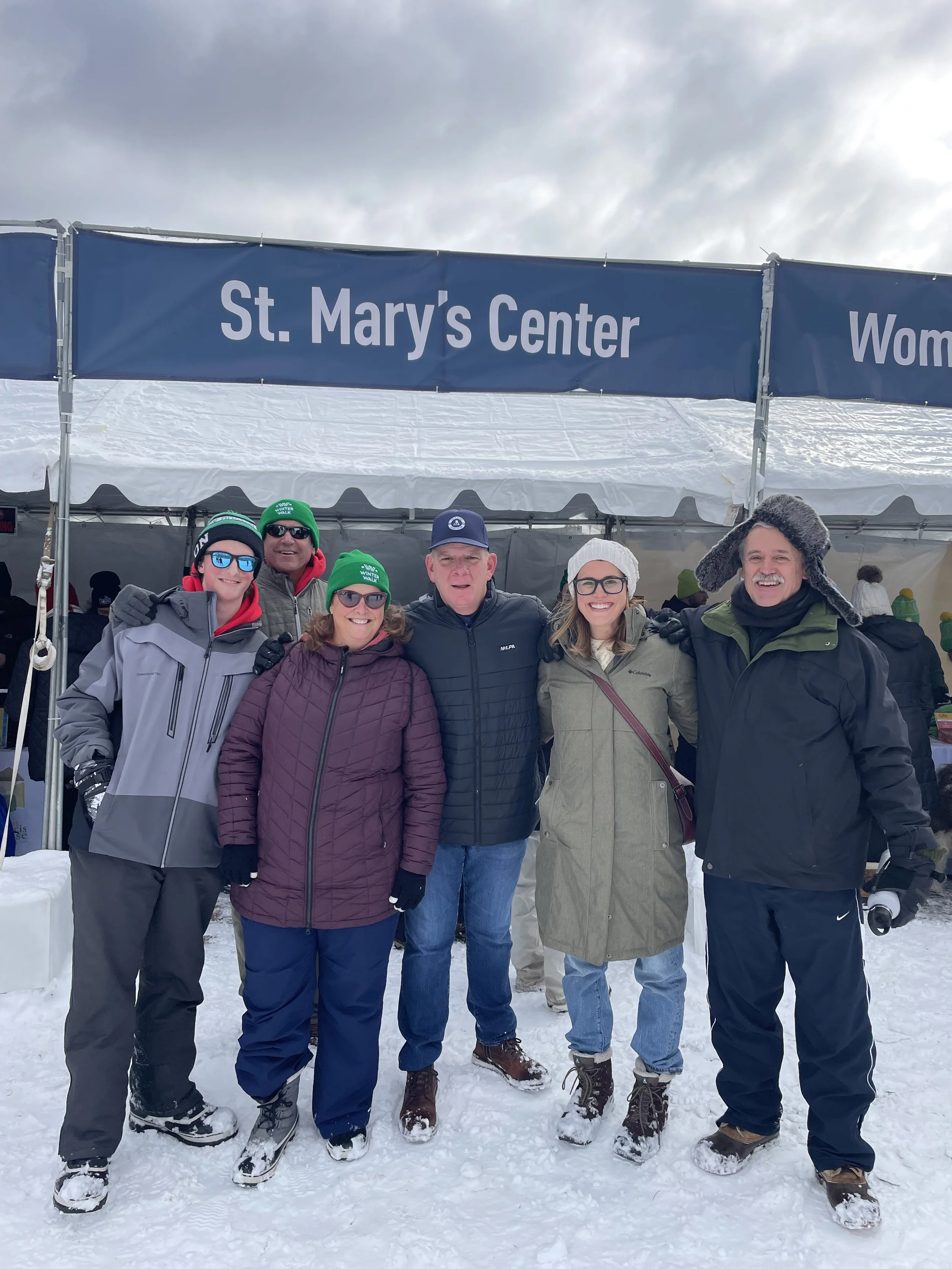6 people gather in their full winter gear in front of the SMC booth at the 2025 winter walk.
