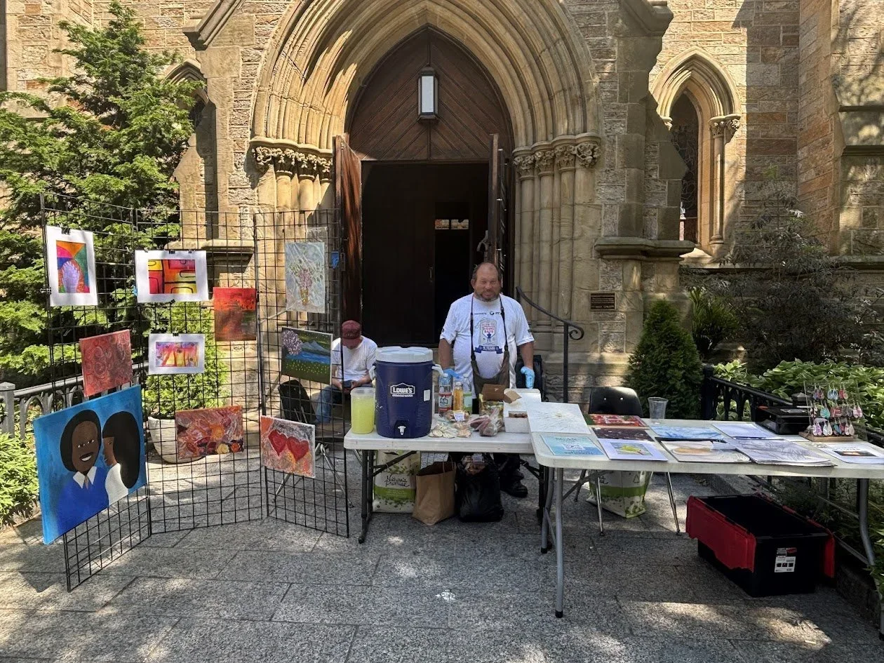 One artist is standing behind two tables and a mesh wire stand displaying the artists artwork. Another man is sitting on the steps right behind him.