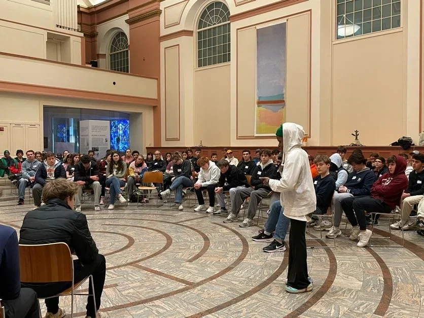 A group of over 50 people gather in a circle with one person standing at the center and speaking.