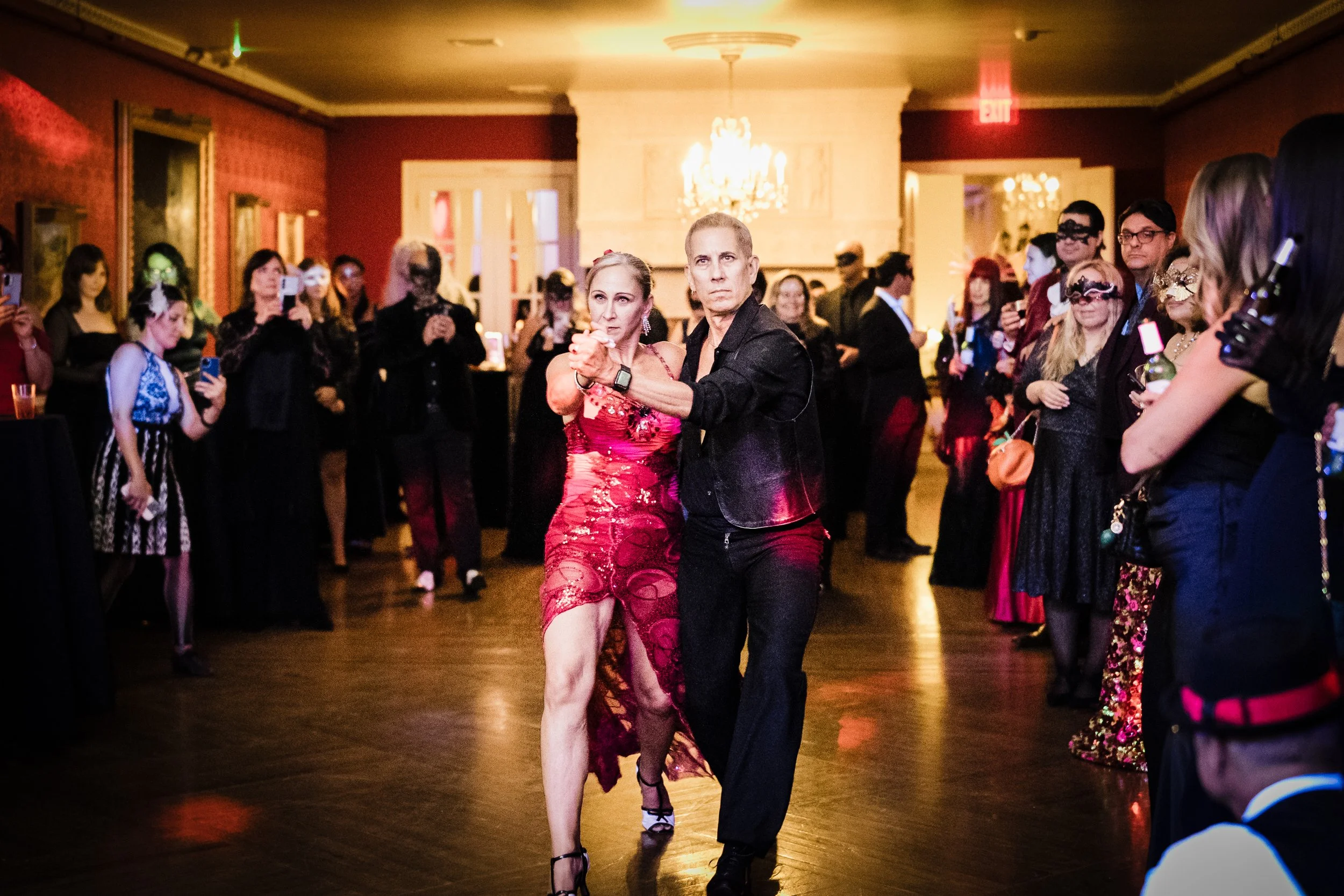 A couple dance closely at a formal event with a crowd watching, some with masks, in an elegant room with chandeliers.