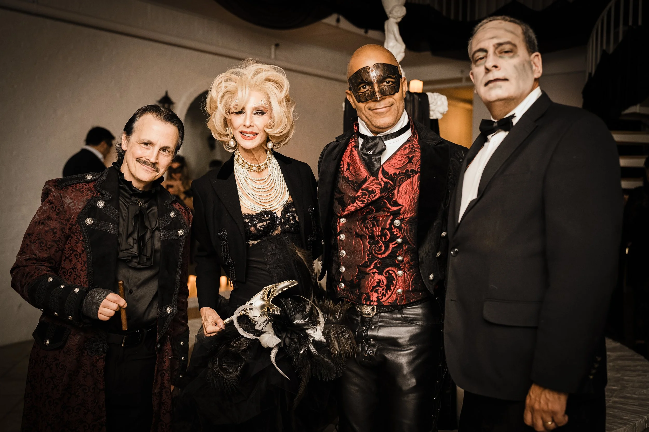 Four people dressed in elegant costumes at a party, including a woman in a black dress with pearls, a man in a mask and red vest, and two men in tuxedos.