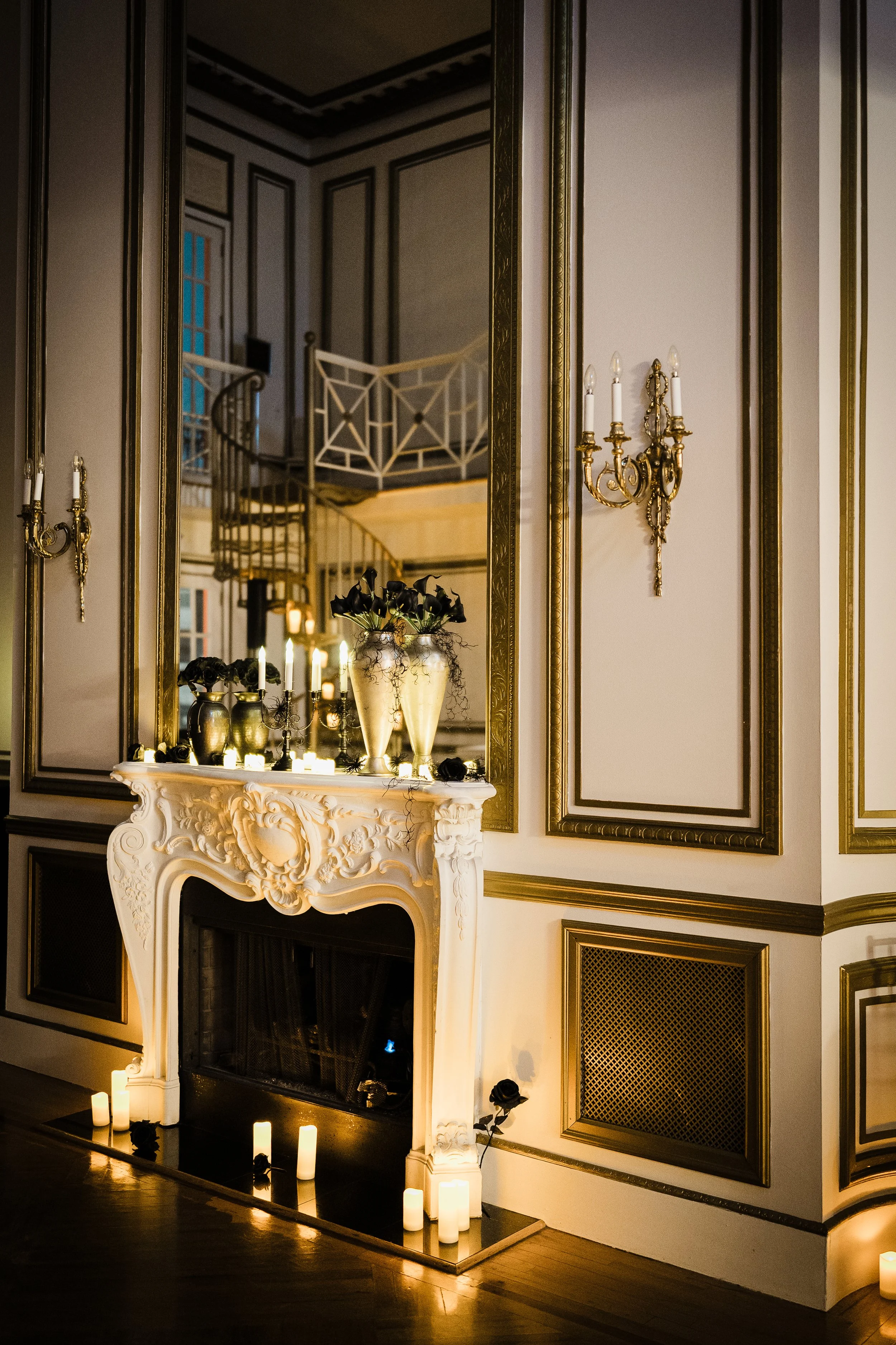 A decorated fireplace mantel with floral arrangements, candles, and black roses, set against a wall adorned with ornate molding and gold accents, with candles placed on the floor in front of the fireplace.