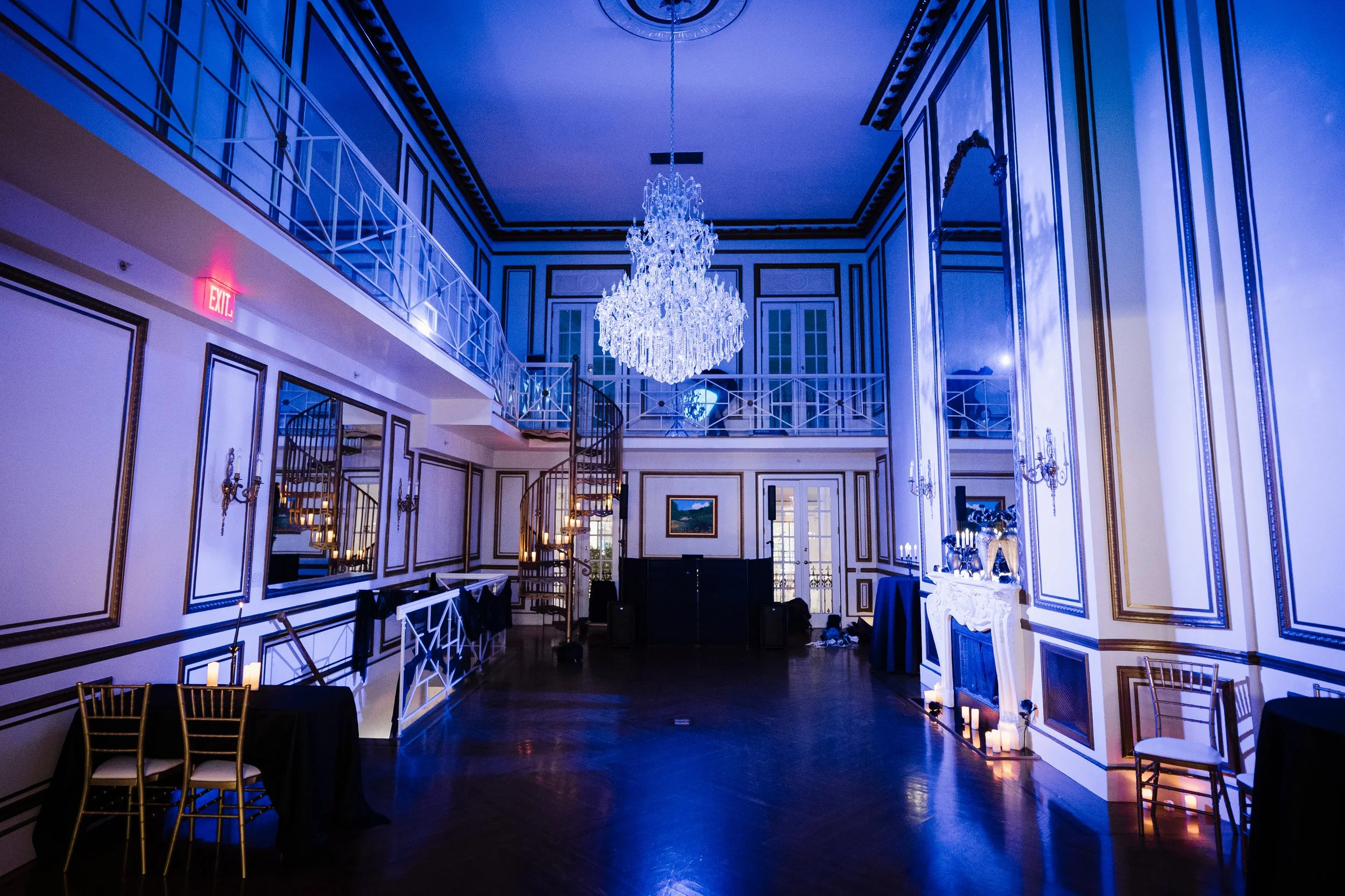 Elegant ballroom with a large crystal chandelier hanging from the ceiling, decorated with white and gold accents, tall windows, and a staircase with white railings, set up for a formal event with chairs and tables.