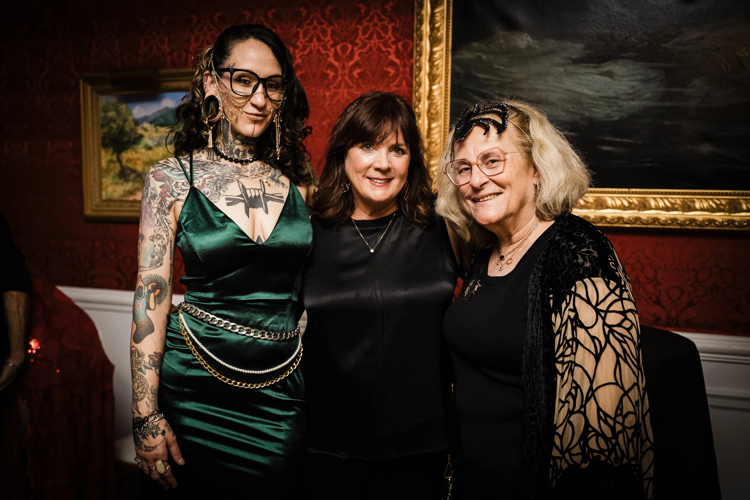 Three women standing together at a social event in a dimly lit room with red patterned wallpaper and framed paintings in the background, smiling at the camera.