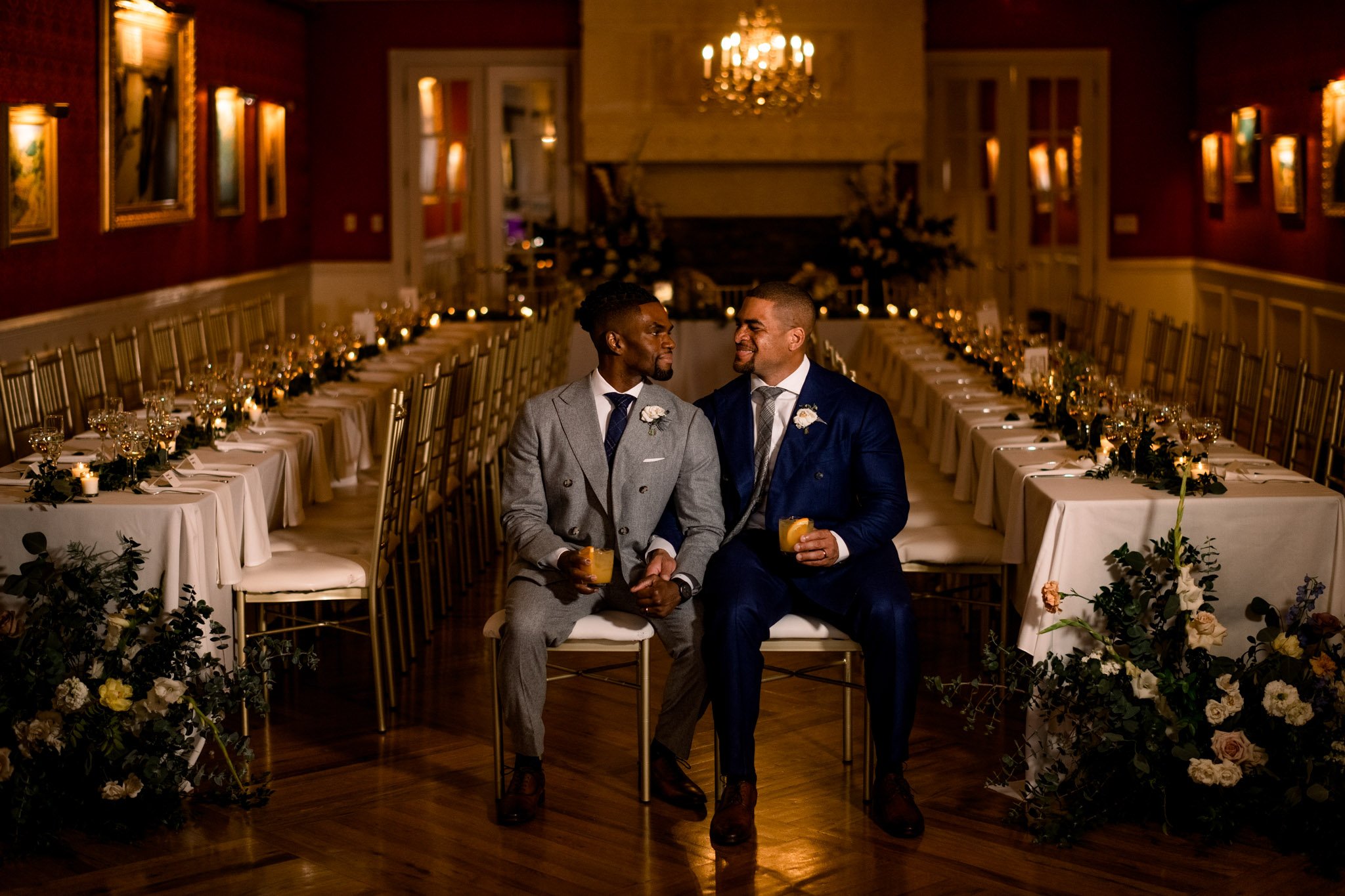 Two men in suits sitting together in a decorated dining hall, smiling at each other, holding drinks, with long banquet tables and floral arrangements.