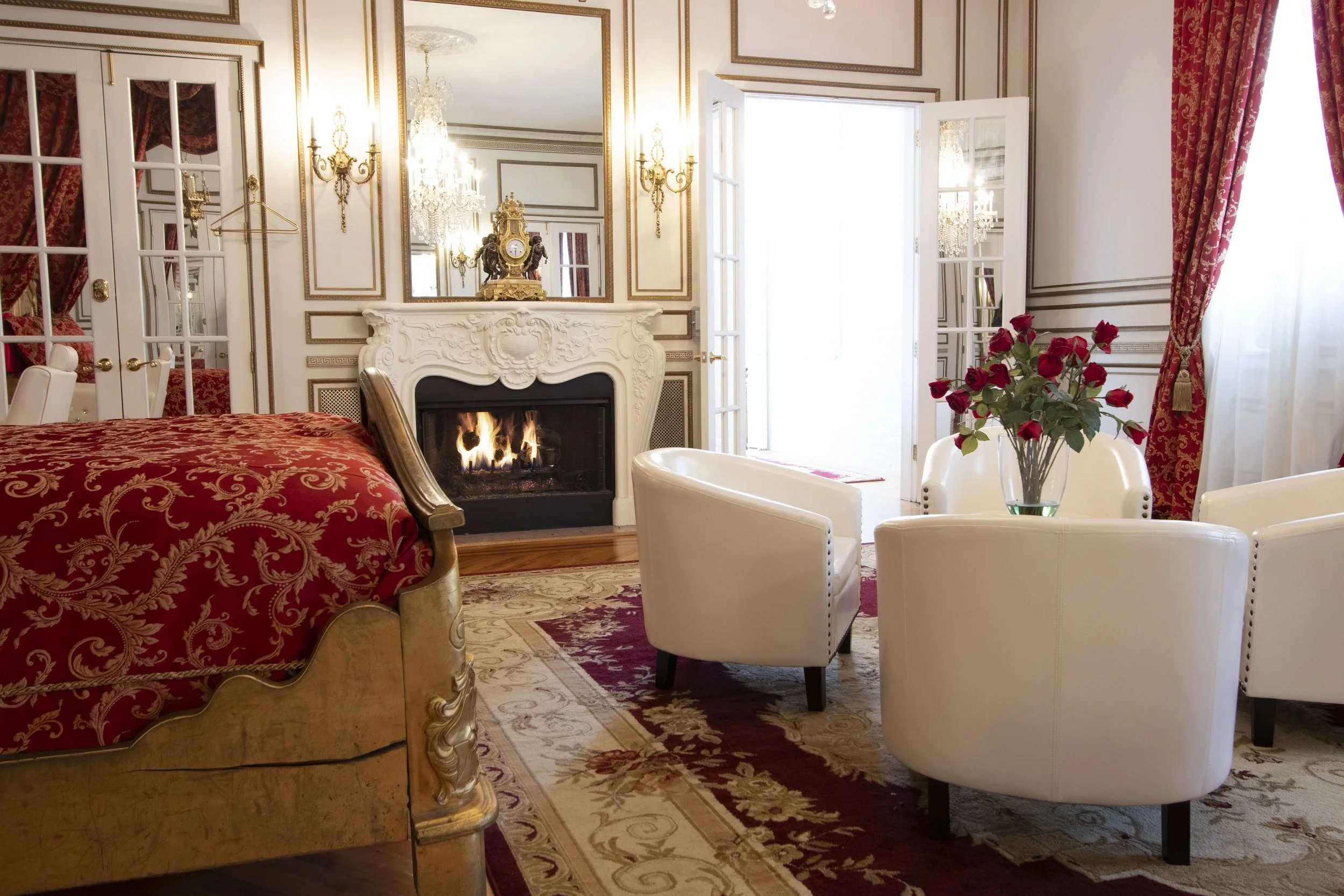 Elegant living room with cream-colored walls, ornate gold trim, a fireplace with a portrait on the mantel, and a large mirror above it. French doors with red and gold drapes lead outside. There is a bouquet of red roses on a table and white armchairs