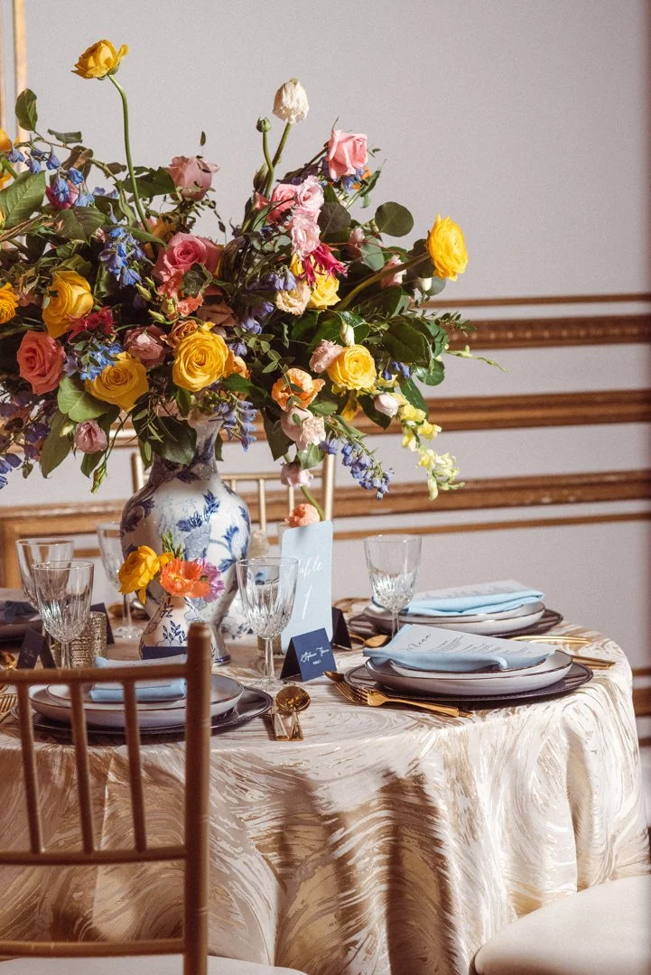 A round table set for a special occasion with a large floral centerpiece in a blue and white vase, gold and crystal glassware, white plates with gold accents, and elegant tableware on a cream-colored tablecloth.
