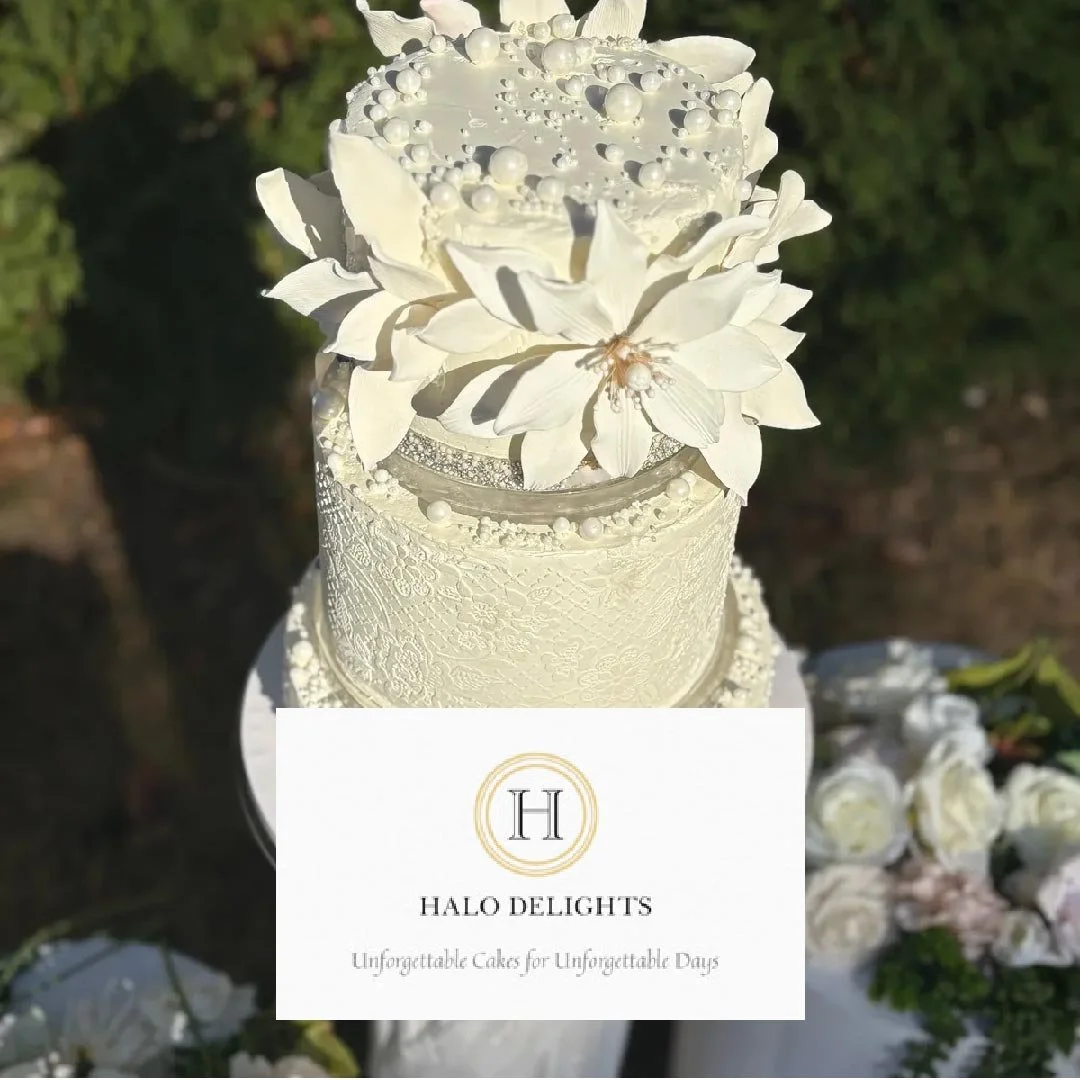 A white cake decorated with white flowers and pearls, with a cake topper resembling a flower arrangement, placed outdoors with a floral arrangement nearby.