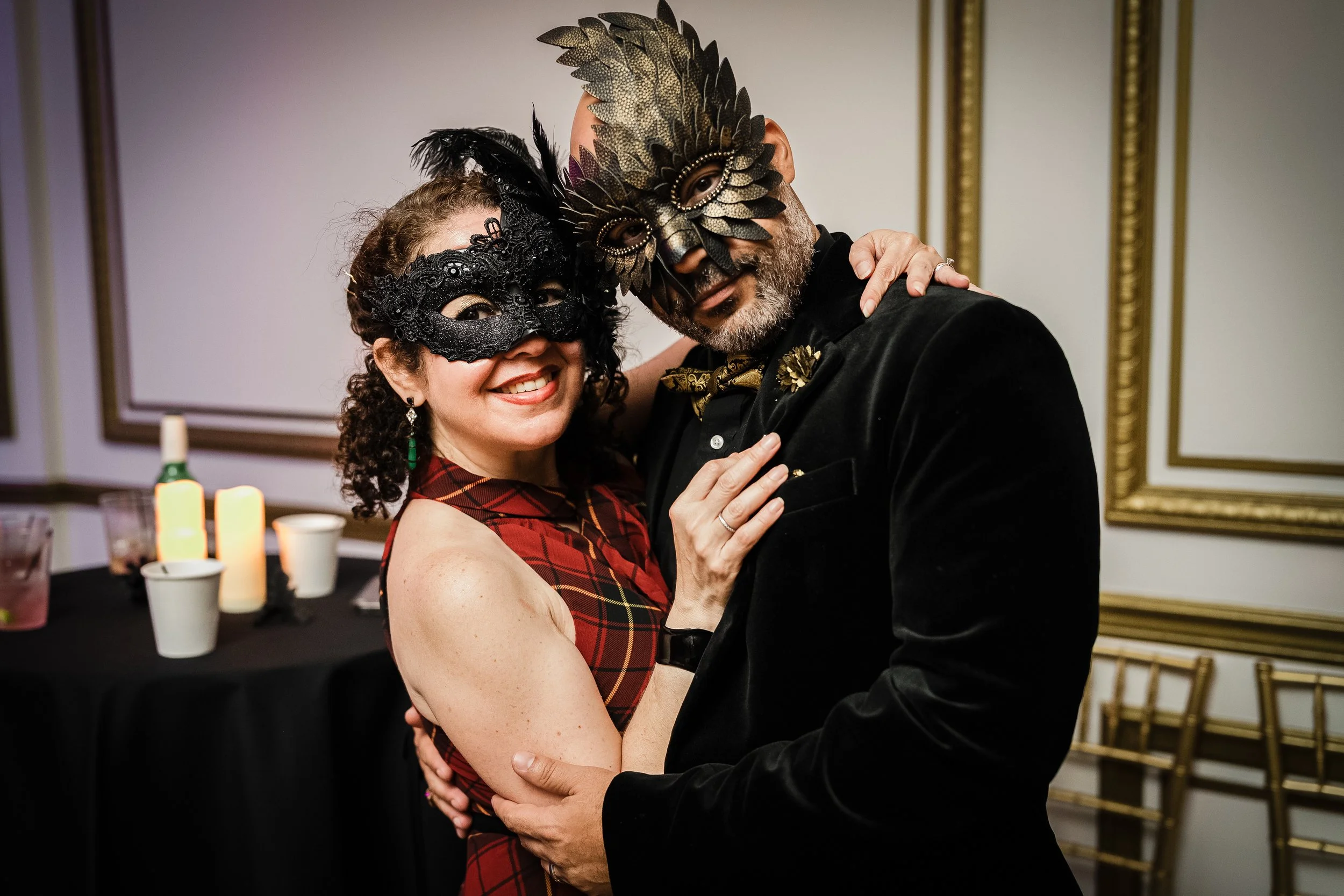 A man and woman wear decorative masks and dance closely at a party or celebration, smiling and holding each other.