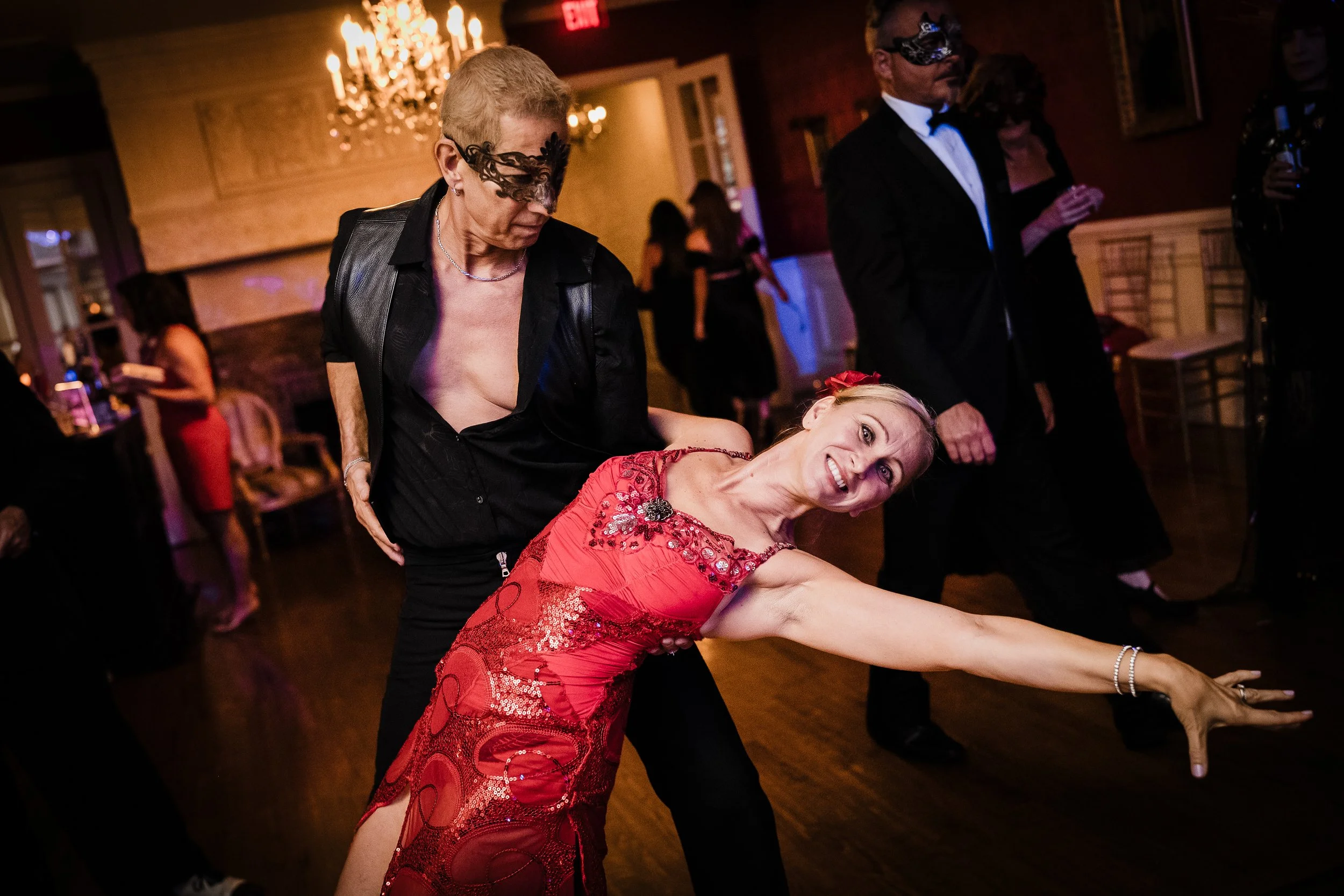 Two women wearing masks are dancing in a dimly lit room with chandeliers. One woman in a black leather jacket is holding and dancing with another woman in a red dress who is smiling.