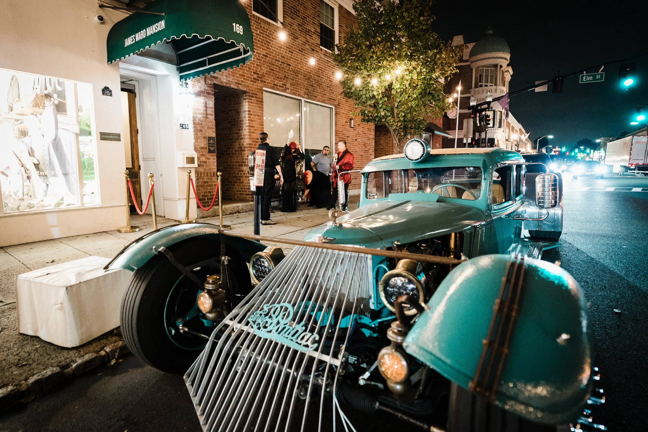 A vintage green car parked on a city street at night, with a brick building and pedestrians visible in the background. The car is in front of a store with a green awning that reads 'James Ward Mansion,' and a group of people are gathered near the ent
