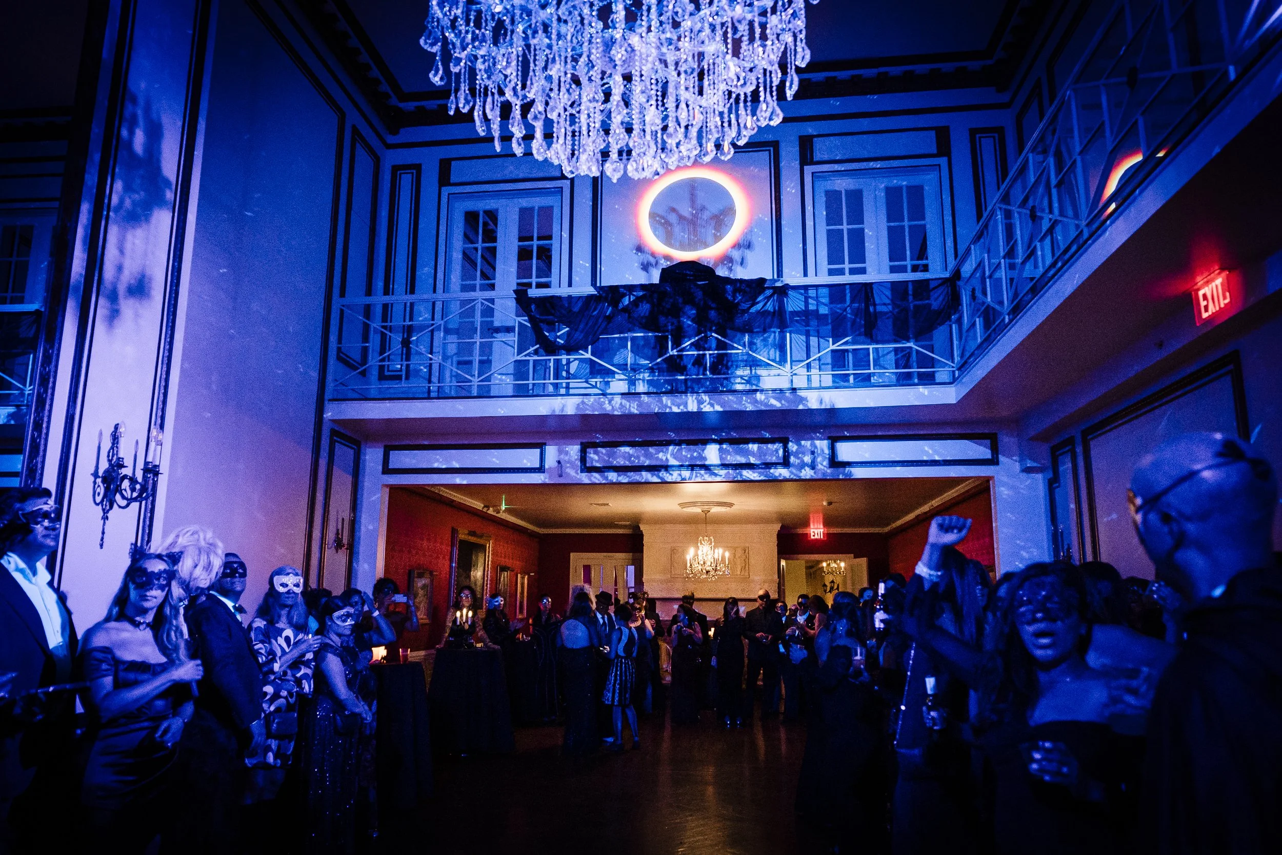 A large indoor party or event with people dressed in formal attire, some wearing masks, standing and raising their fists. The room features a chandelier hanging from the ceiling, with blue lighting and a projected ring and tree design on the wall. Th