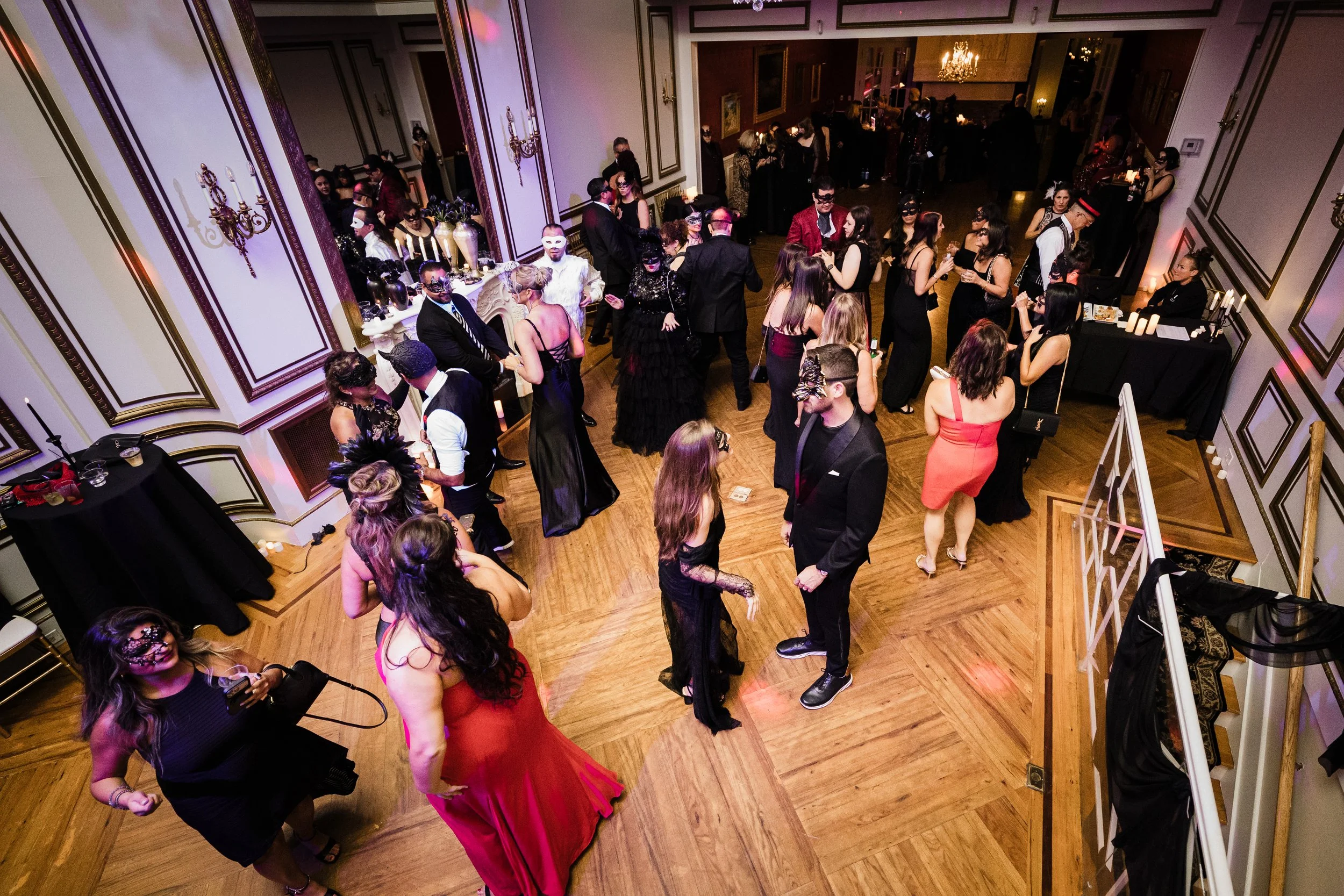 Guests at a masquerade ball dancing and socializing in a luxurious ballroom with ornate wall moldings, chandeliers, and a table with masks and decorations.
