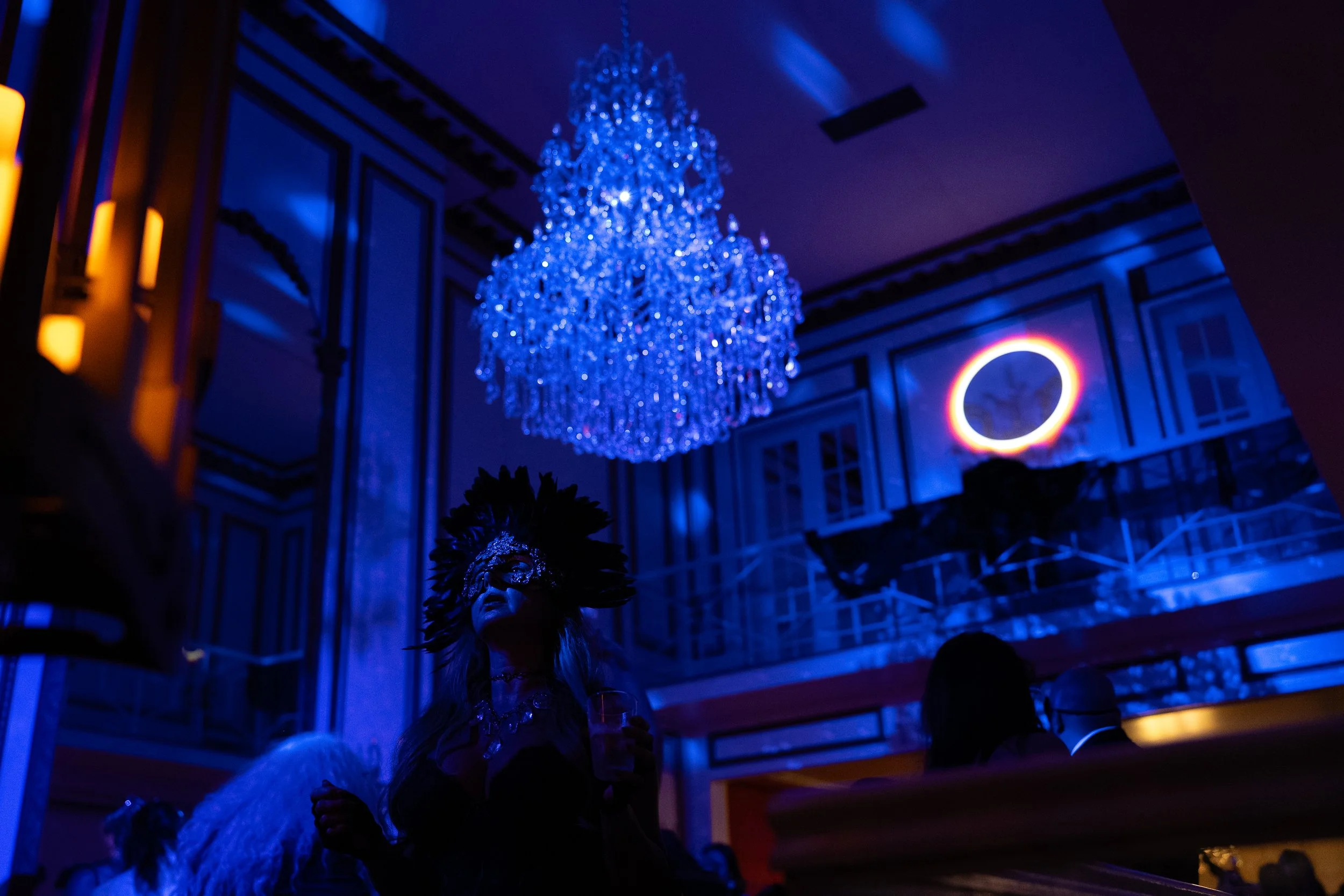 A large, elaborate chandelier lit with bright blue lights hangs from the ceiling of an elegant room with dark wood paneling. There is a person in a masquerade mask and feathered costume in the foreground, suggesting a masquerade or costume event.