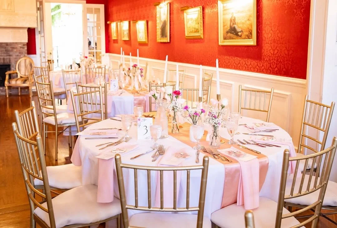 A decorated banquet table set for a celebration in a room with red walls, gold chairs, and framed paintings.