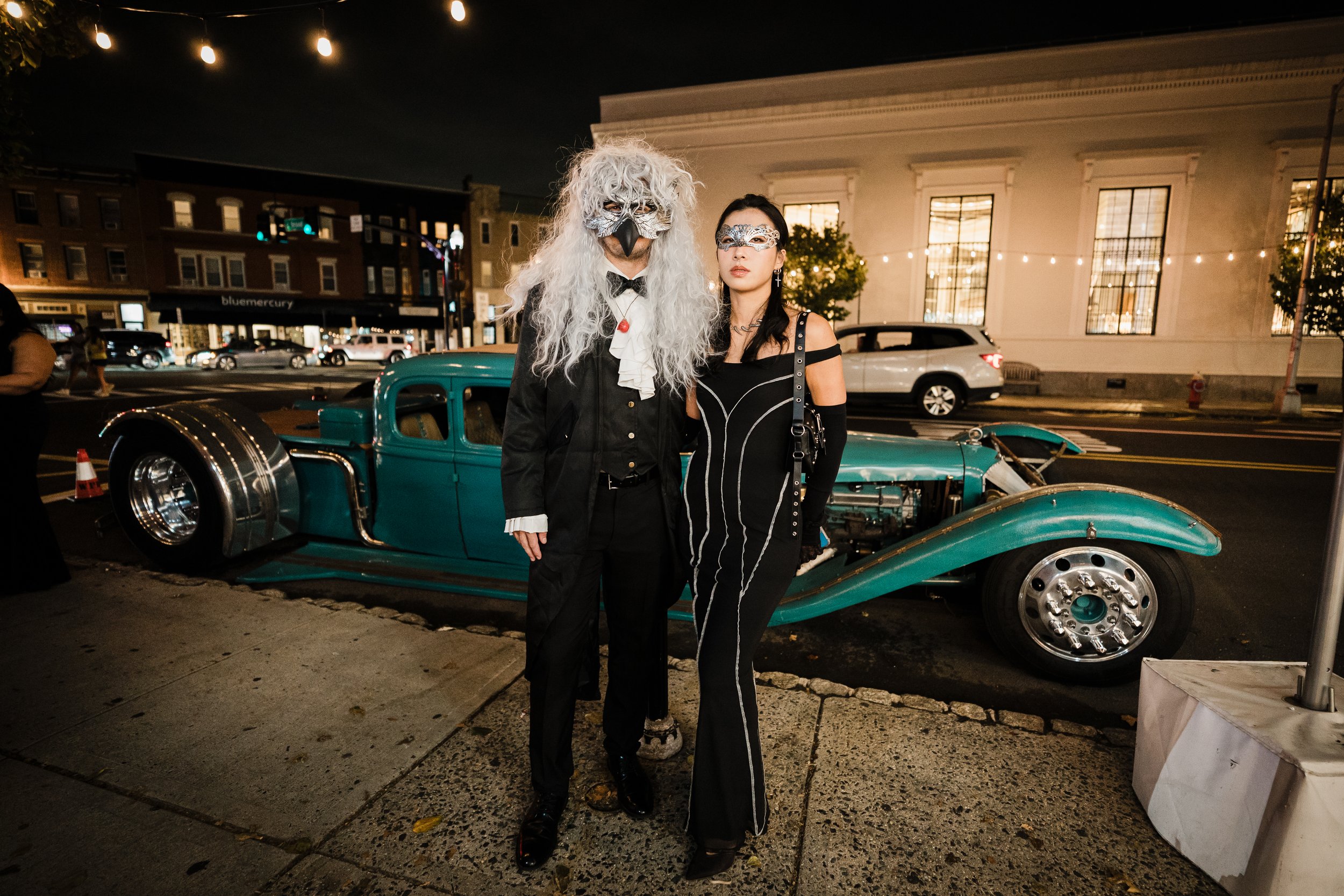 Two women in masquerade masks pose next to a vintage turquoise car at night, with string lights overhead and illuminated buildings in the background.