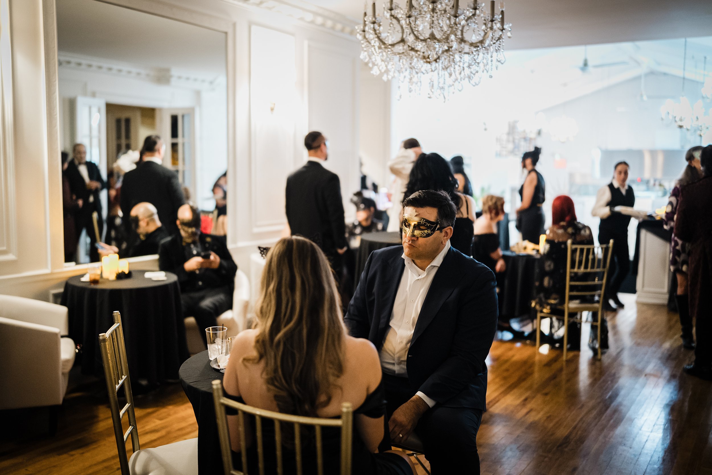 People dressed in formal attire at a social event in an elegant decorated room with a chandelier, some wearing masks, engaging in conversation.