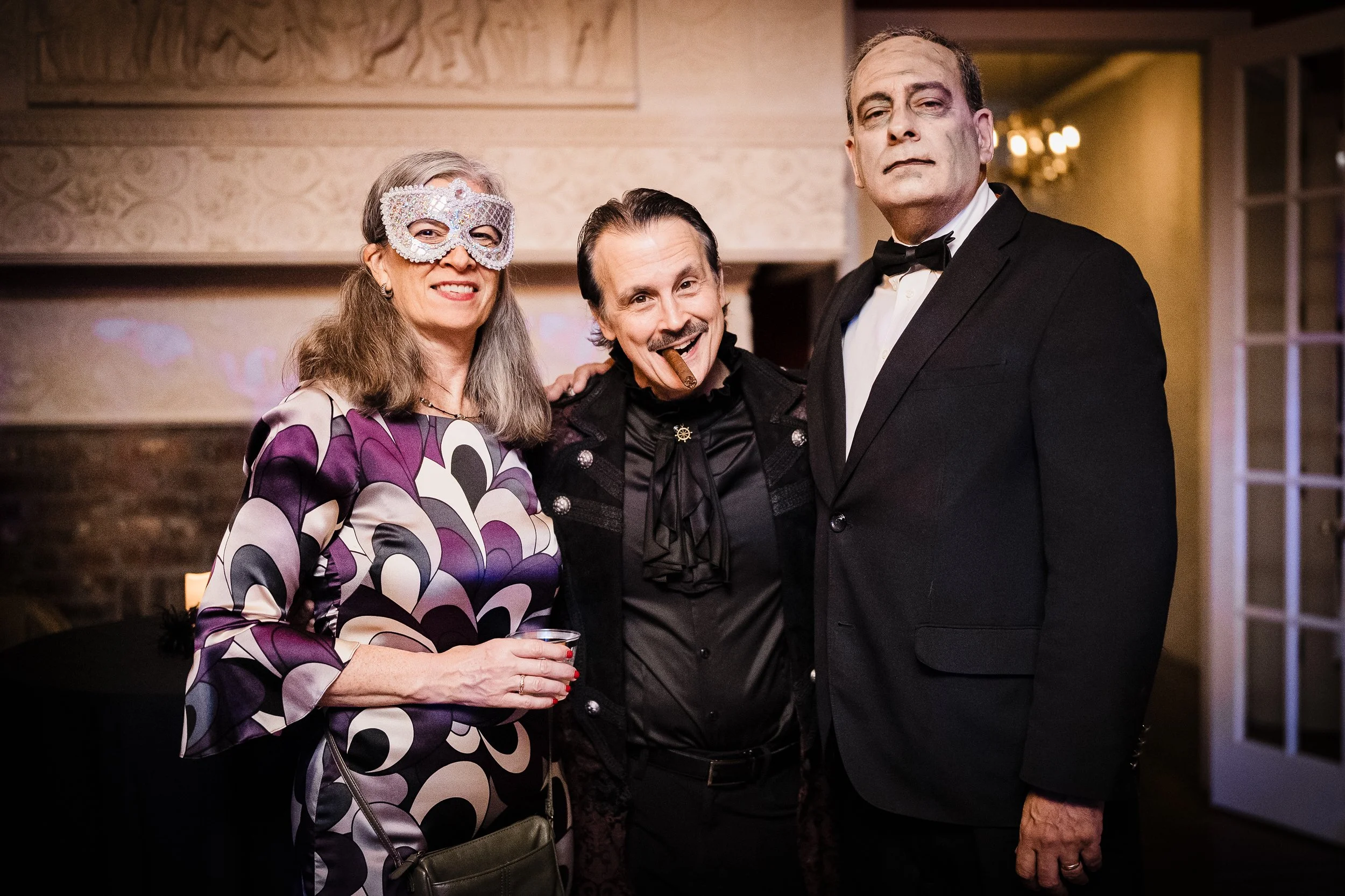 Group of three adults dressed in formal attire at a social event, one woman wearing a decorative mask and a woman with a cigar, standing indoors with warm lighting.