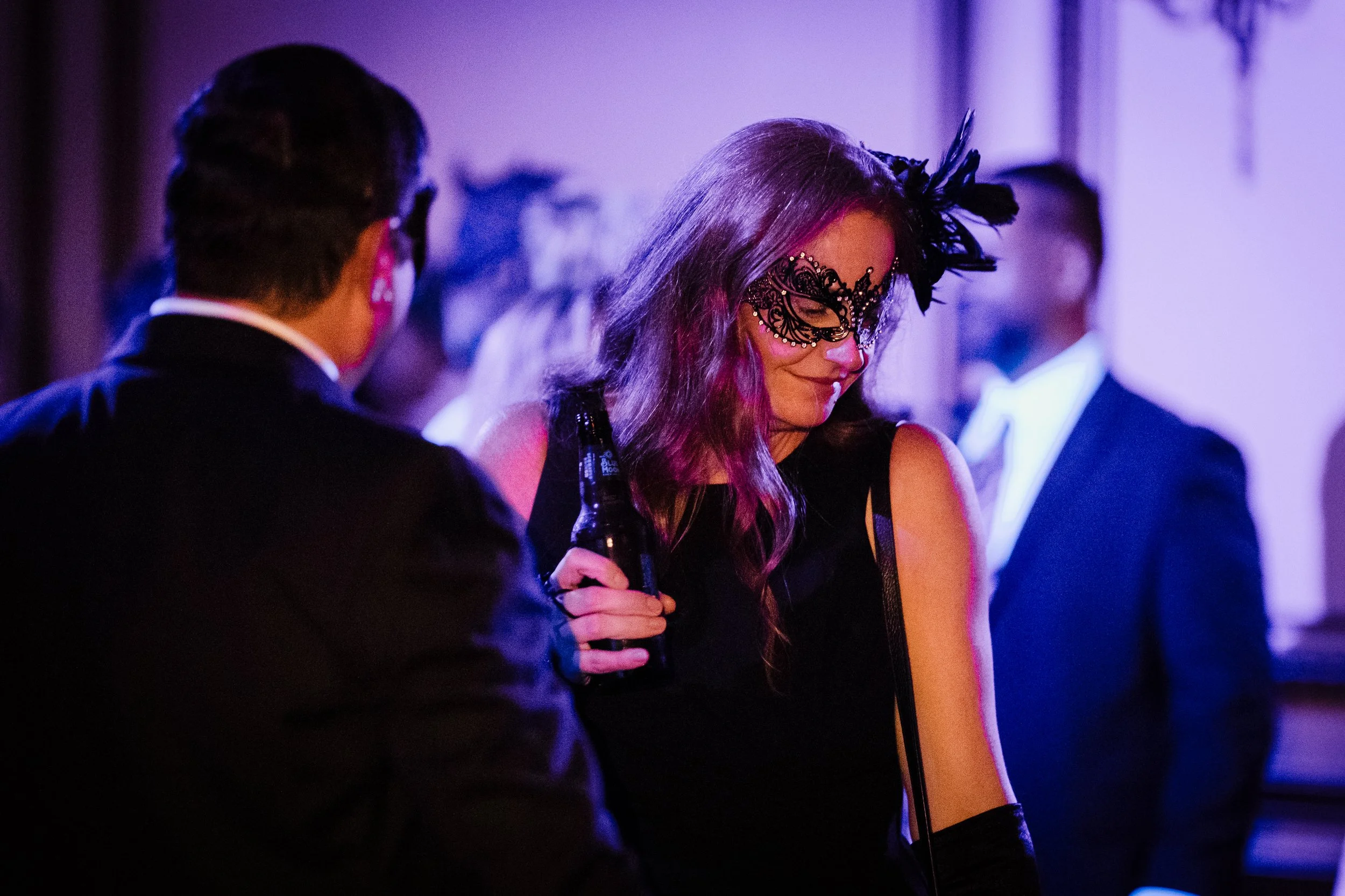 A woman wearing a black lace mask with feathers and a black dress, holding a bottle, at a party with purple lighting. Two men in tuxedos are present, one facing her and the other in the background.