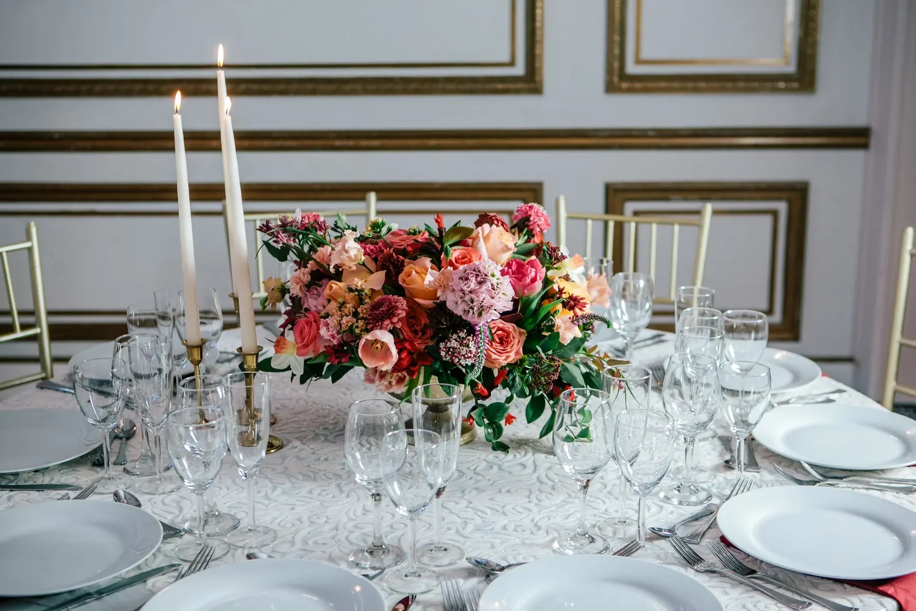 Elegant dining table decorated with a large colorful floral centerpiece, surrounded by clear glassware, white plates, and silver cutlery, set against a decorated wall with gold framing.