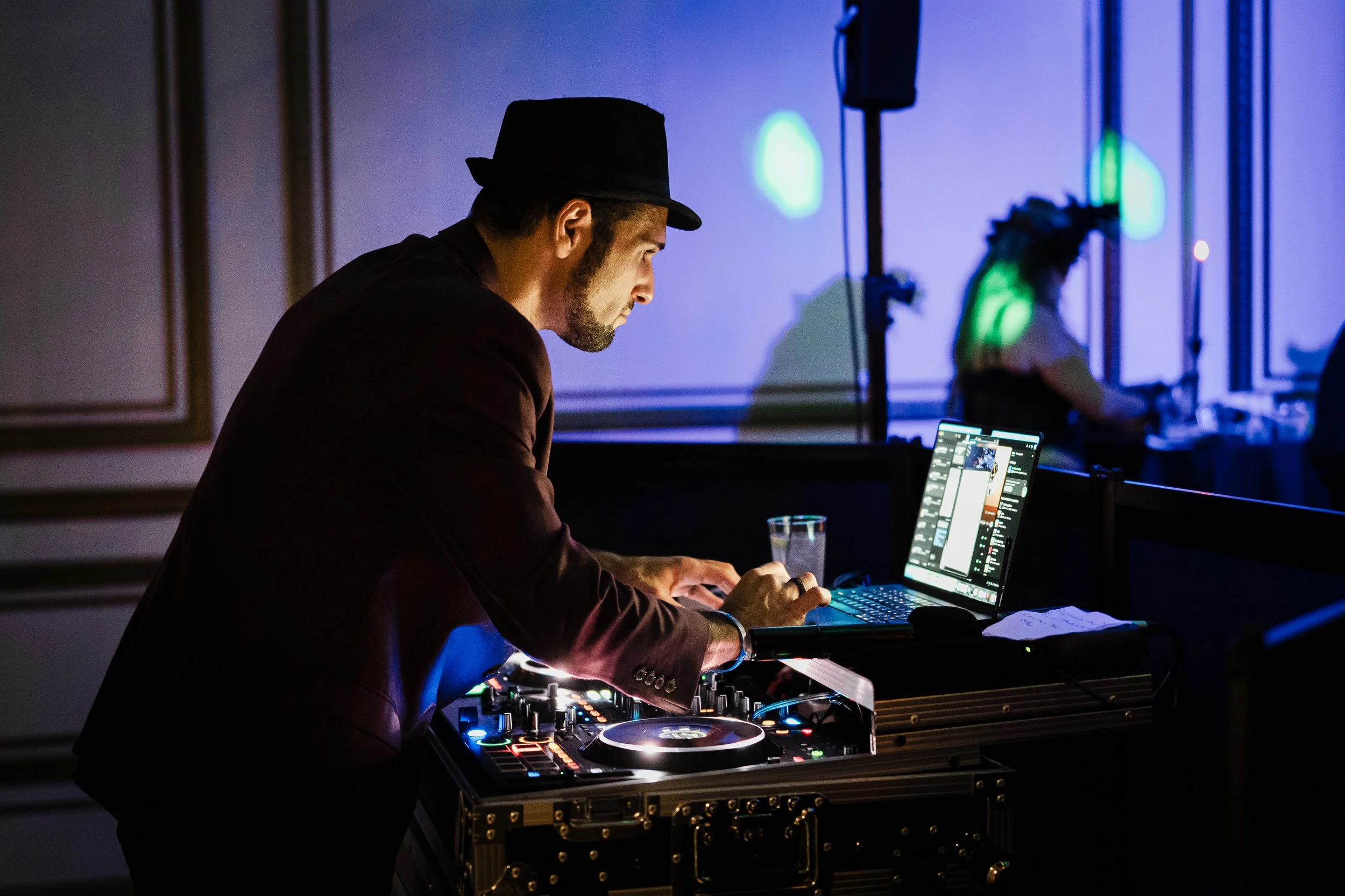 A DJ wearing a black hat and brown suit, focused on his equipment, performing at a dimly lit event, with a person with green hair in the background.