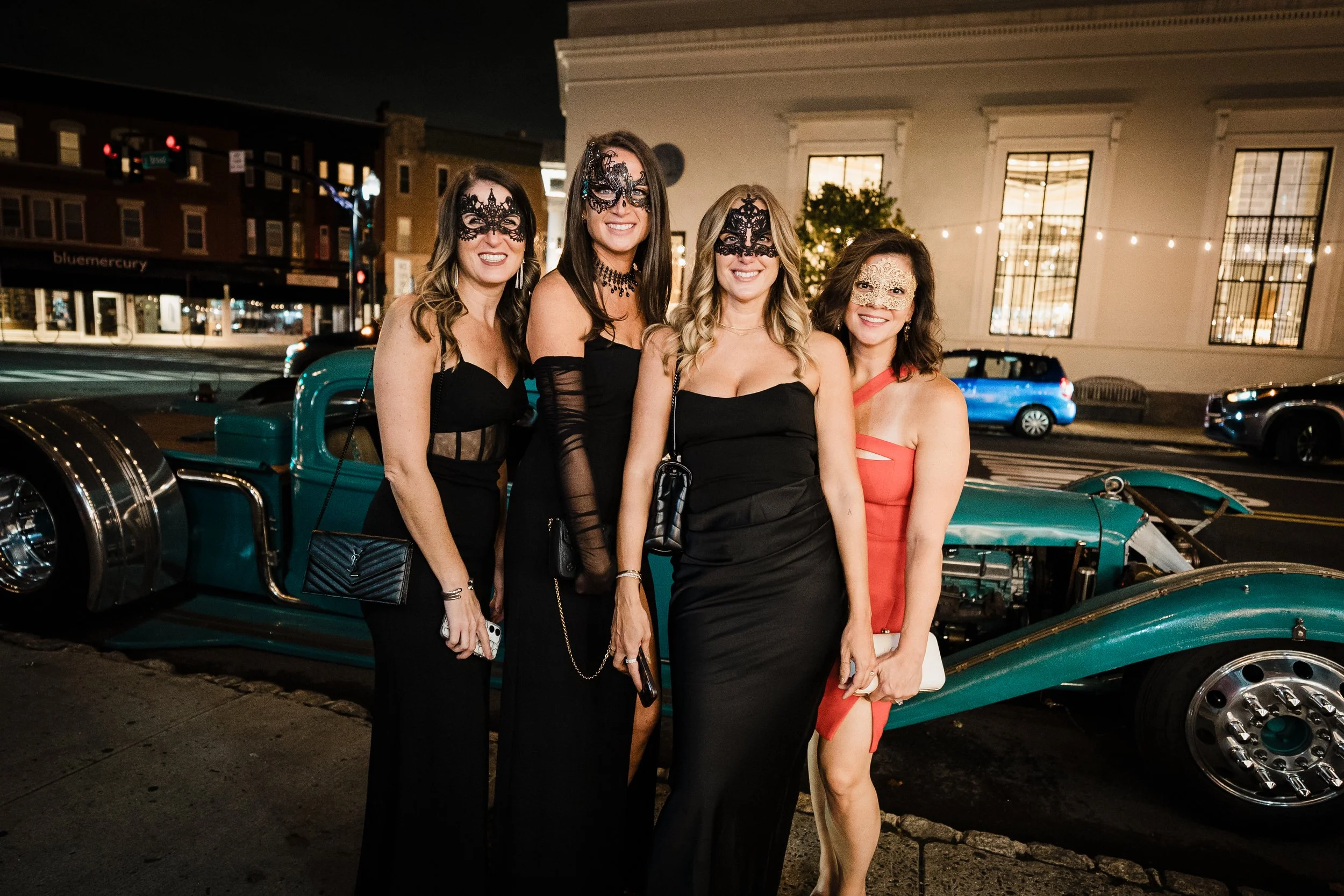 Four women dressed in black evening gowns with lace masks, standing in front of a vintage green car on a city street at night, celebrating at an event.