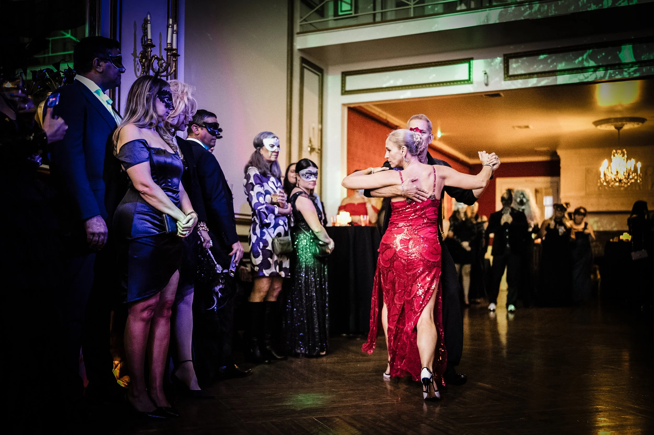 A woman in a red gown is dancing with a man in a black suit at a party while a line of people wearing masquerade masks watch. The background has elegant decor and warm lighting.