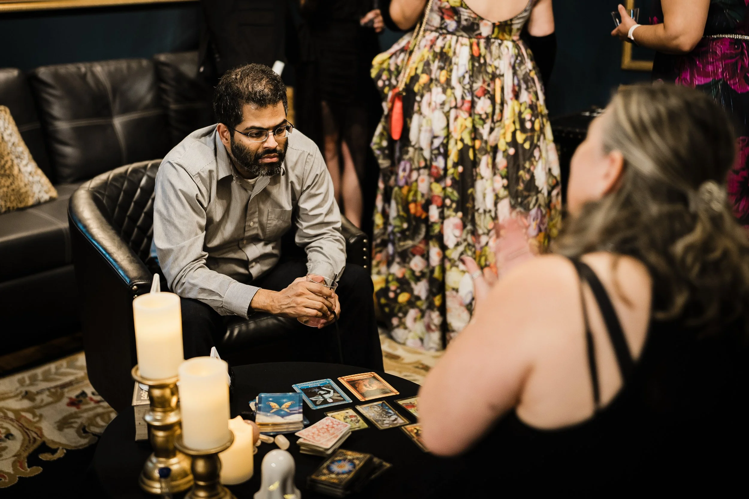 A man with glasses and a beard sitting on a black chair in front of a table with tarot cards, candles, and decorative items, surrounded by women standing and sitting in a dimly lit room, possibly engaging in a tarot reading or spiritual gathering.