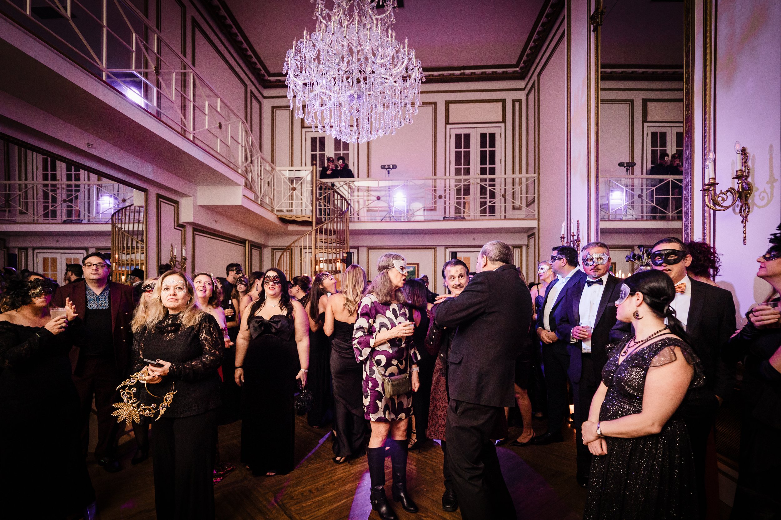 People dressed in formal attire and wearing masks at a masquerade ball in an elegant, chandelier-lit ballroom.