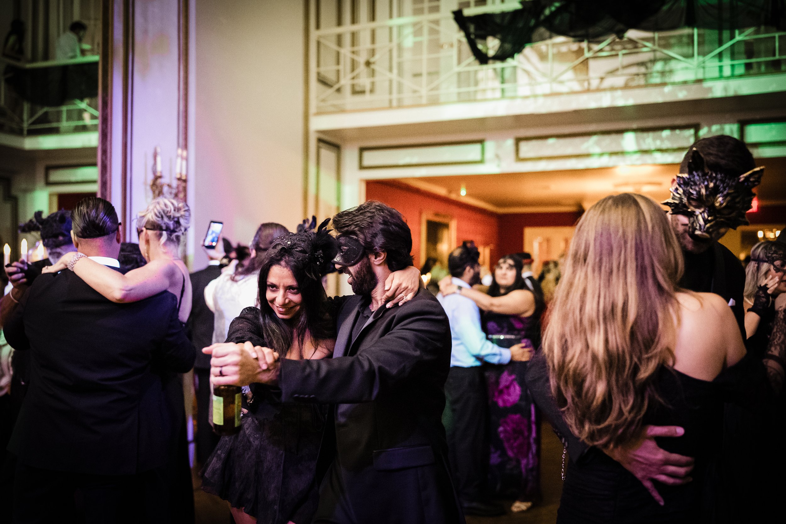 People dressed in formal attire dancing at a masquerade ball, with masks and costumes, in a decorated indoor venue.