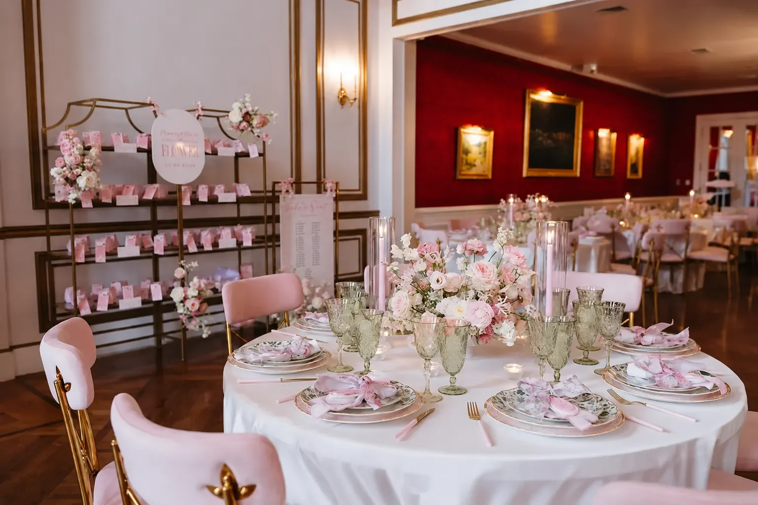Elegant round table set for a wedding reception with pink and white floral centerpieces, gold-rimmed plates, pink napkins, and gold flatware, in a decorated banquet hall.