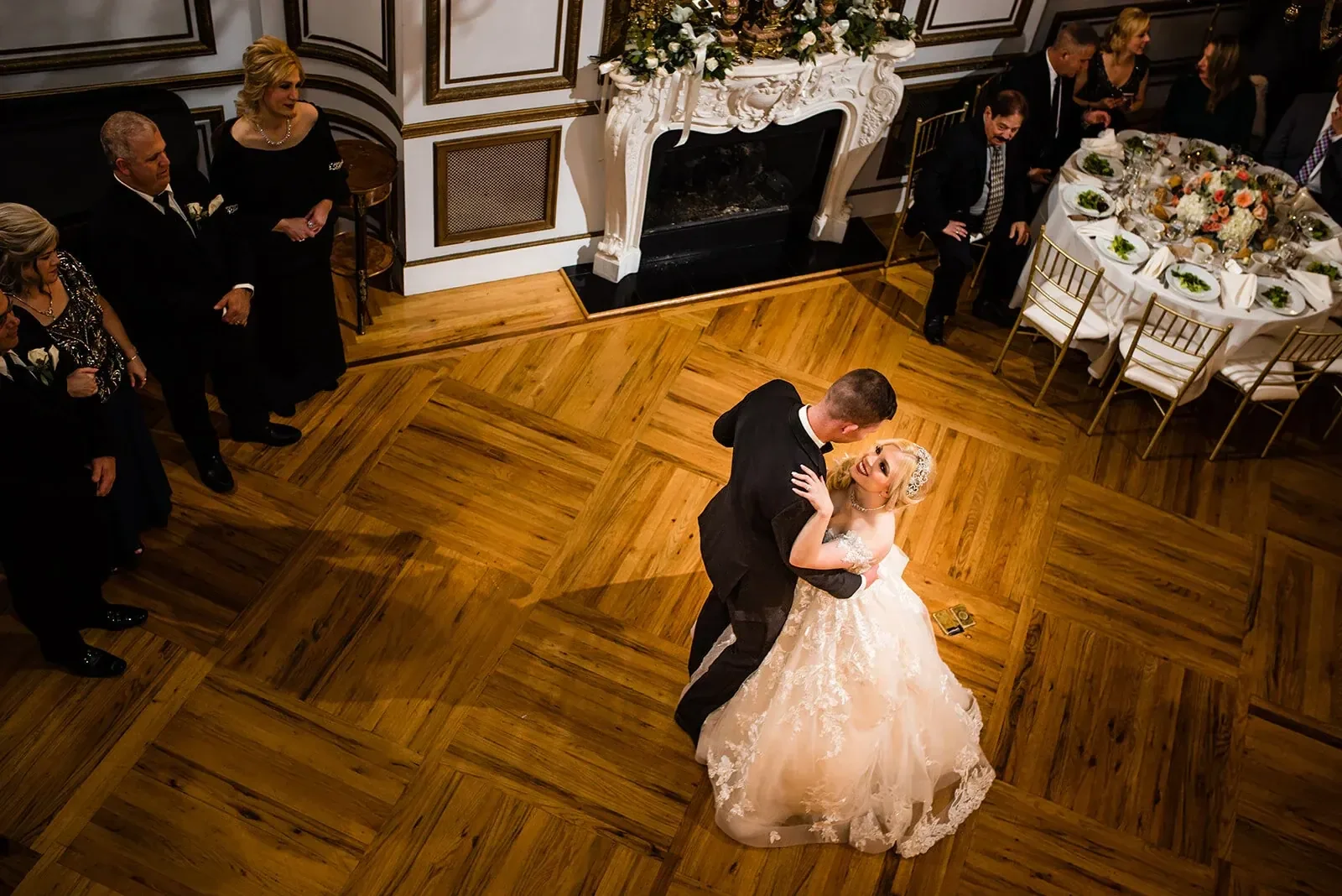 A bride and groom dancing at their wedding reception, surrounded by seated guests in an elegant, decorated ballroom.