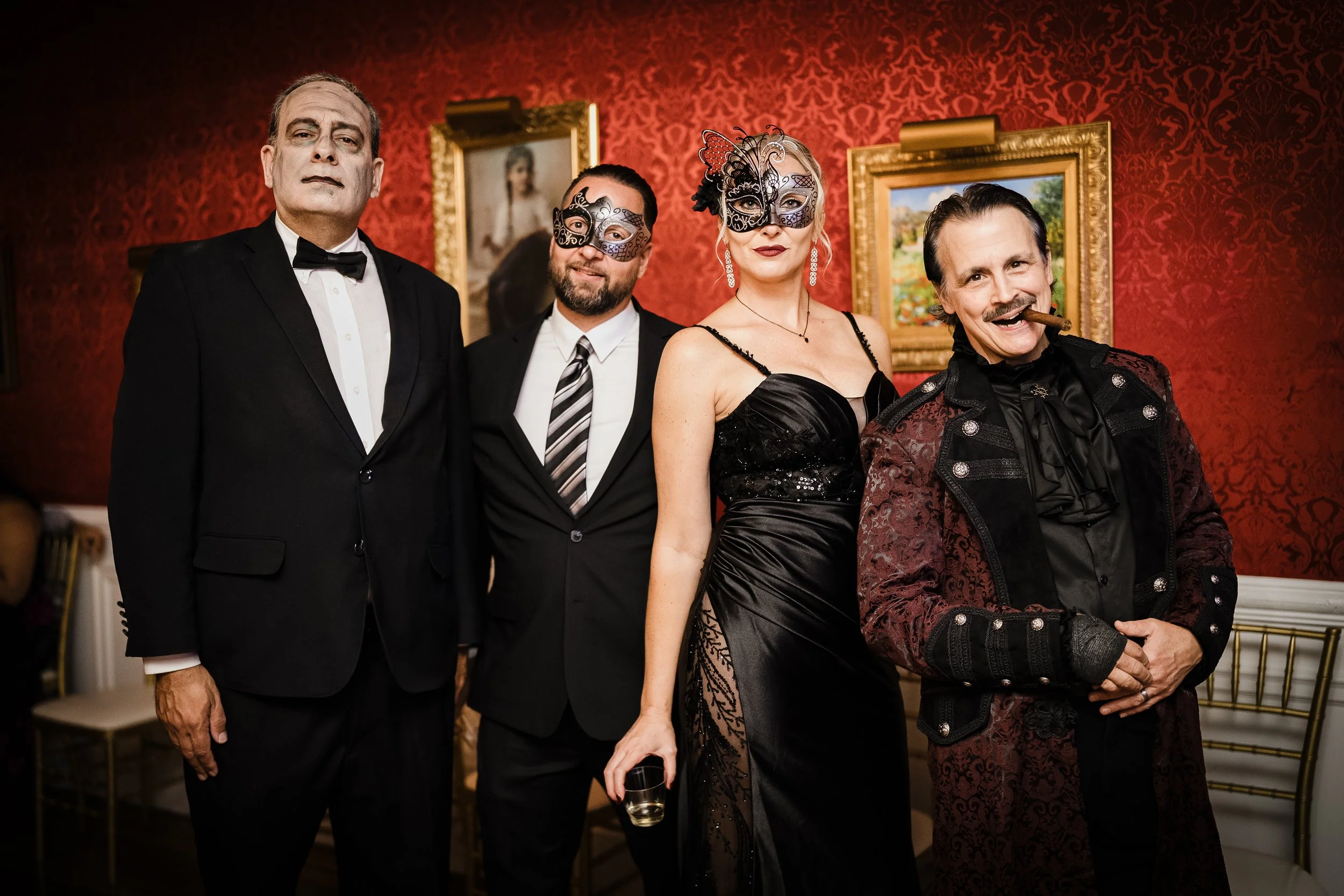 Group of four adults dressed in formal and costume attire at what appears to be a fancy party or event. Two individuals wear masks, and one man has a cigar in his mouth. The background features red patterned wallpaper and framed artwork.