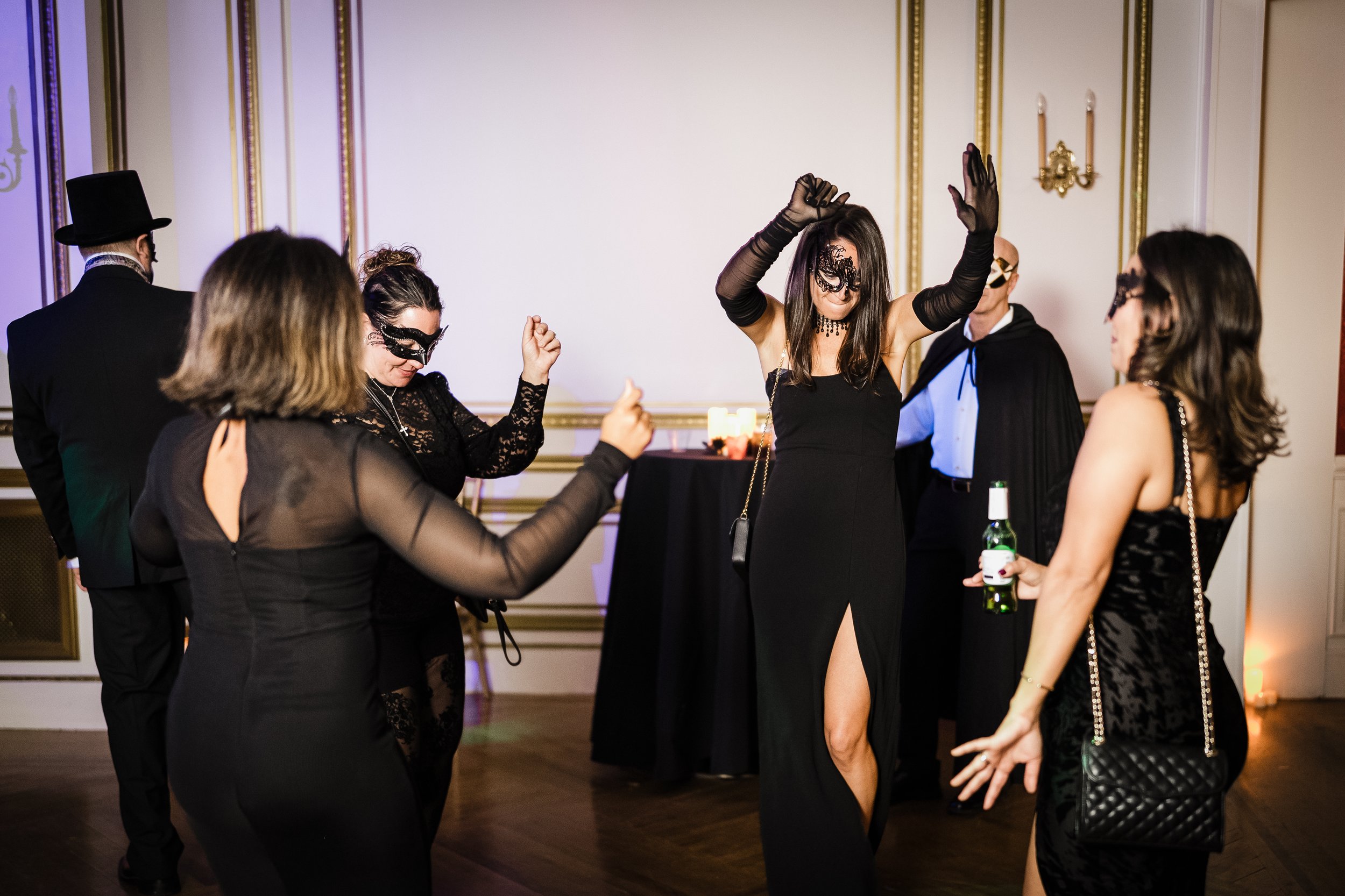 Group of people dressed in fancy attire at a masquerade party, dancing and socializing in a decorated room with gold trim on the walls.