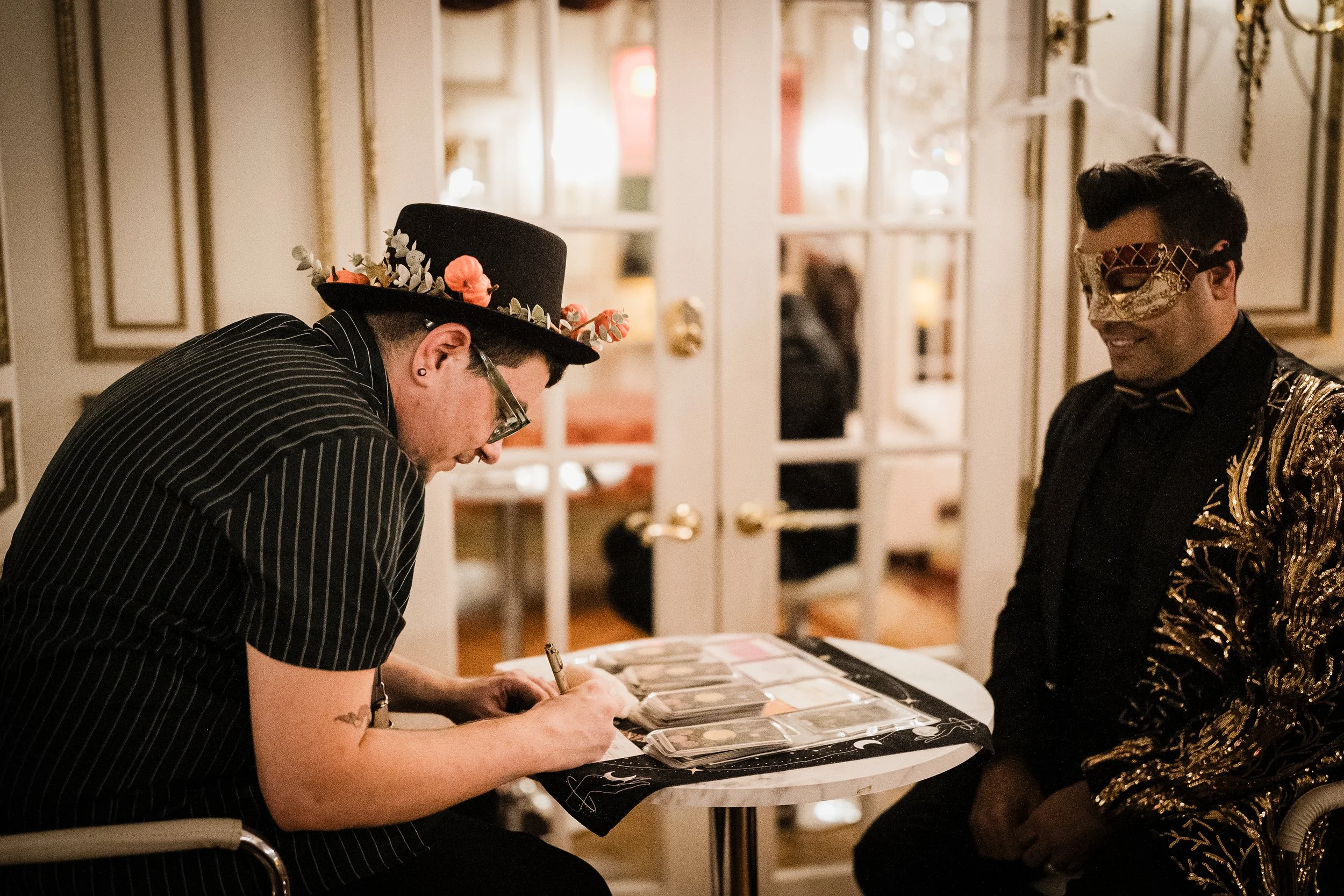 Two men wearing decorative masks and fancy clothing standing at a table with photographs, one signing a photograph, inside a decorated room.