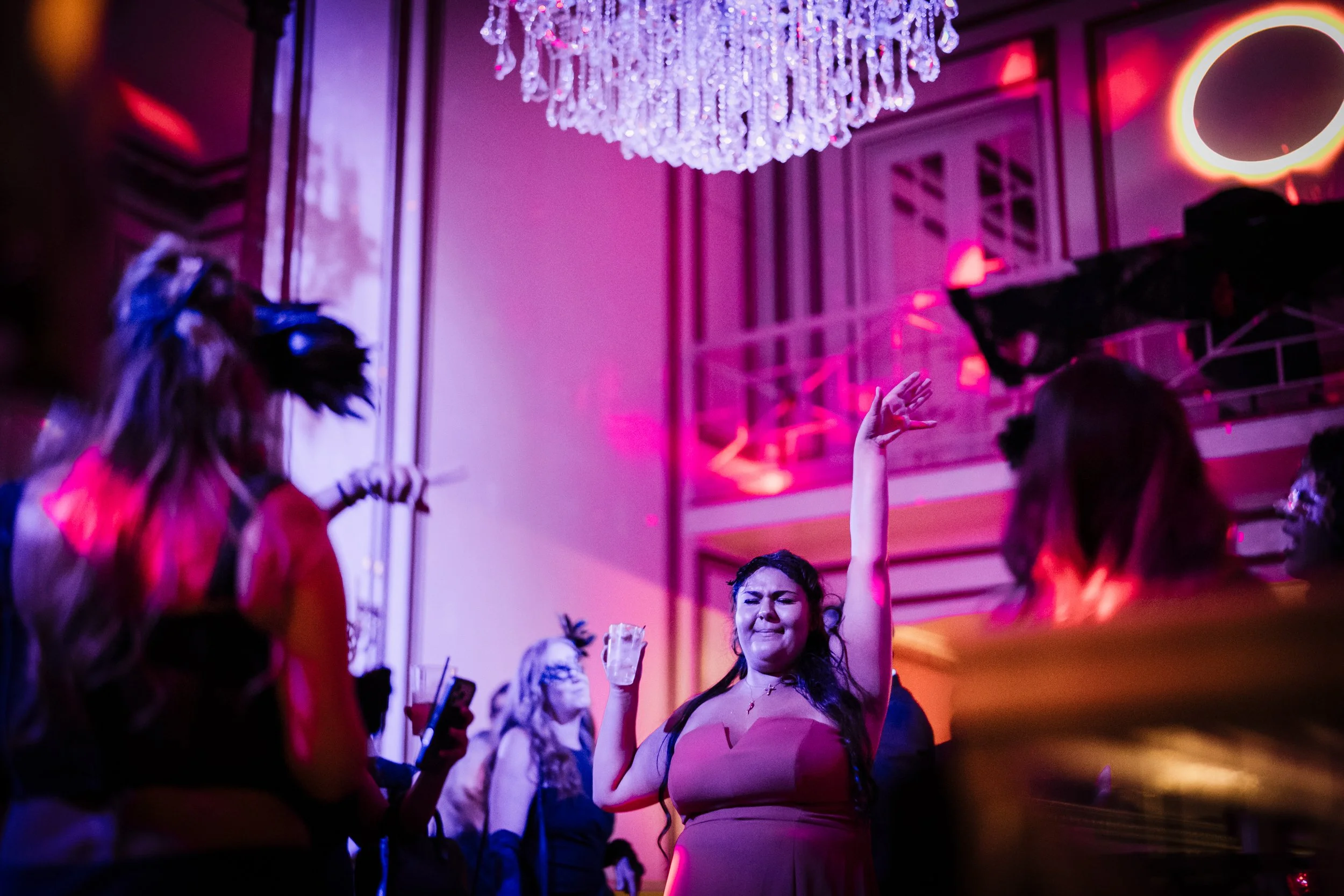 People dancing and socializing at a party with pink and purple lighting, a chandelier, and a large circular decorative mirror on the wall.