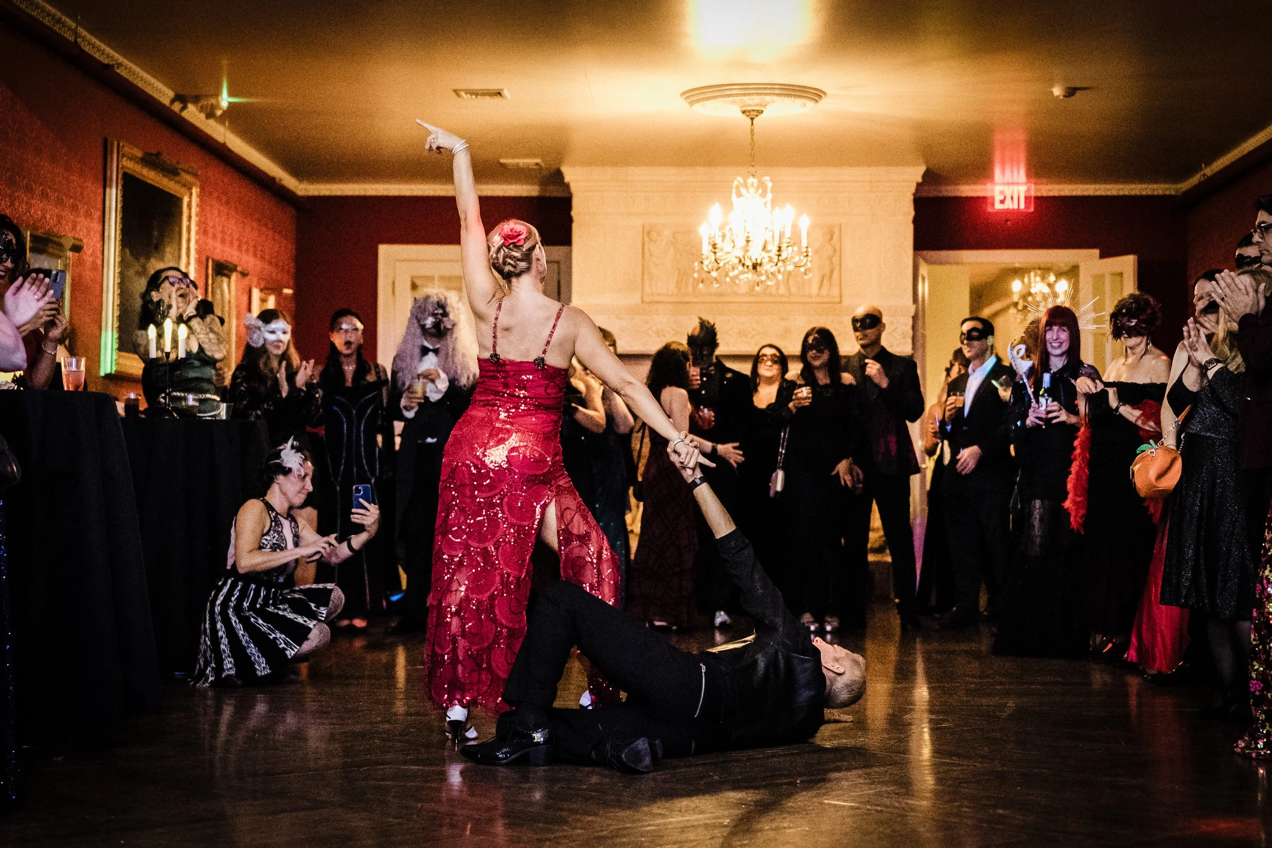 A performer dressed in a red gown with fishnet details dances on stage with a person kneeling underneath her during a lively event with many spectators. The audience appears excited, and some are dressed in costumes, masks, or masks.
