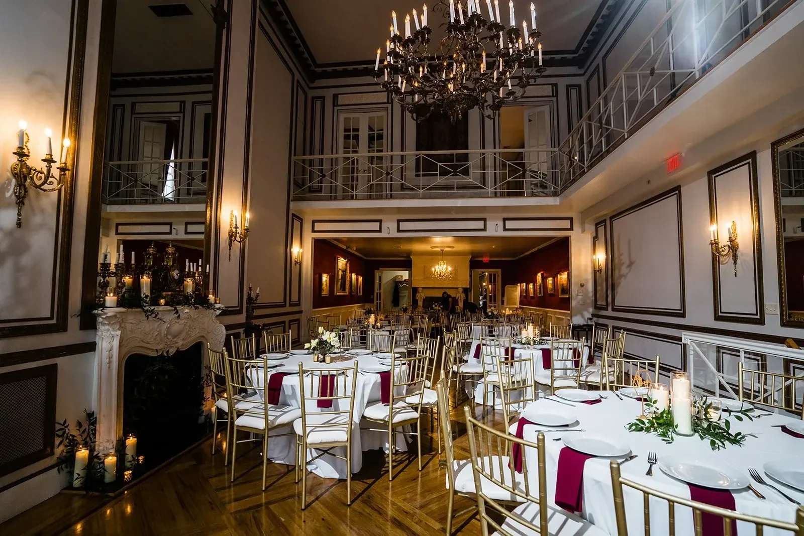 Elegant banquet hall decorated for a formal event with round tables set with white tablecloths, burgundy napkins, candles, and floral centerpieces. The room features gold chairs, a high ceiling with chandeliers, wall sconces, paneled walls, a fireplace with candles, and a second-floor balcony.