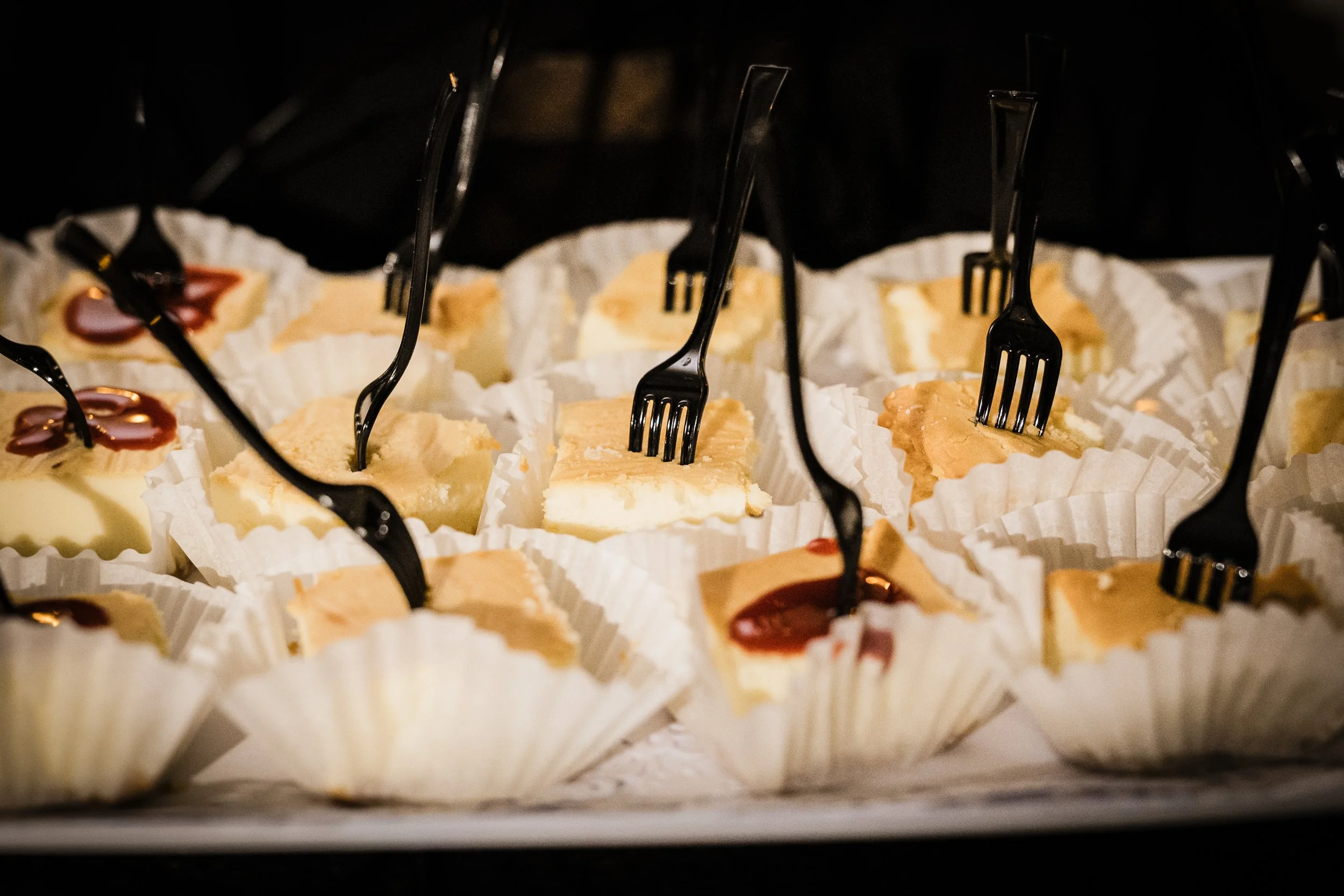 Assorted cheesecake slices in paper cupcake liners, lined up on a baking tray with black forks inserted into each. The background is dark.