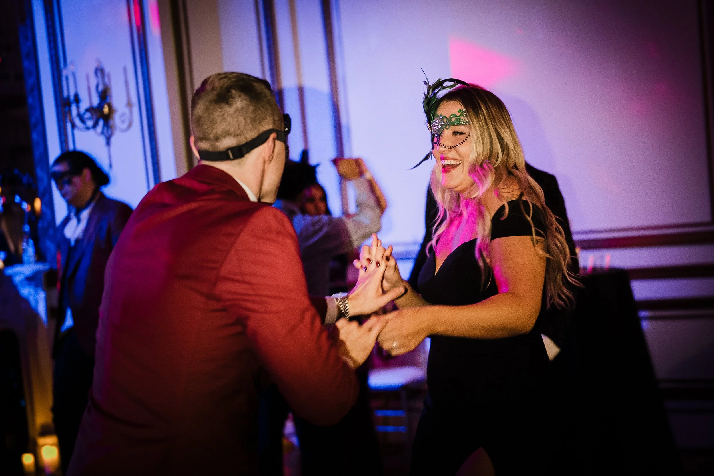 A couple dancing happily at a party, wearing masks and festive attire, with blurred party guests in the background and colorful lighting.