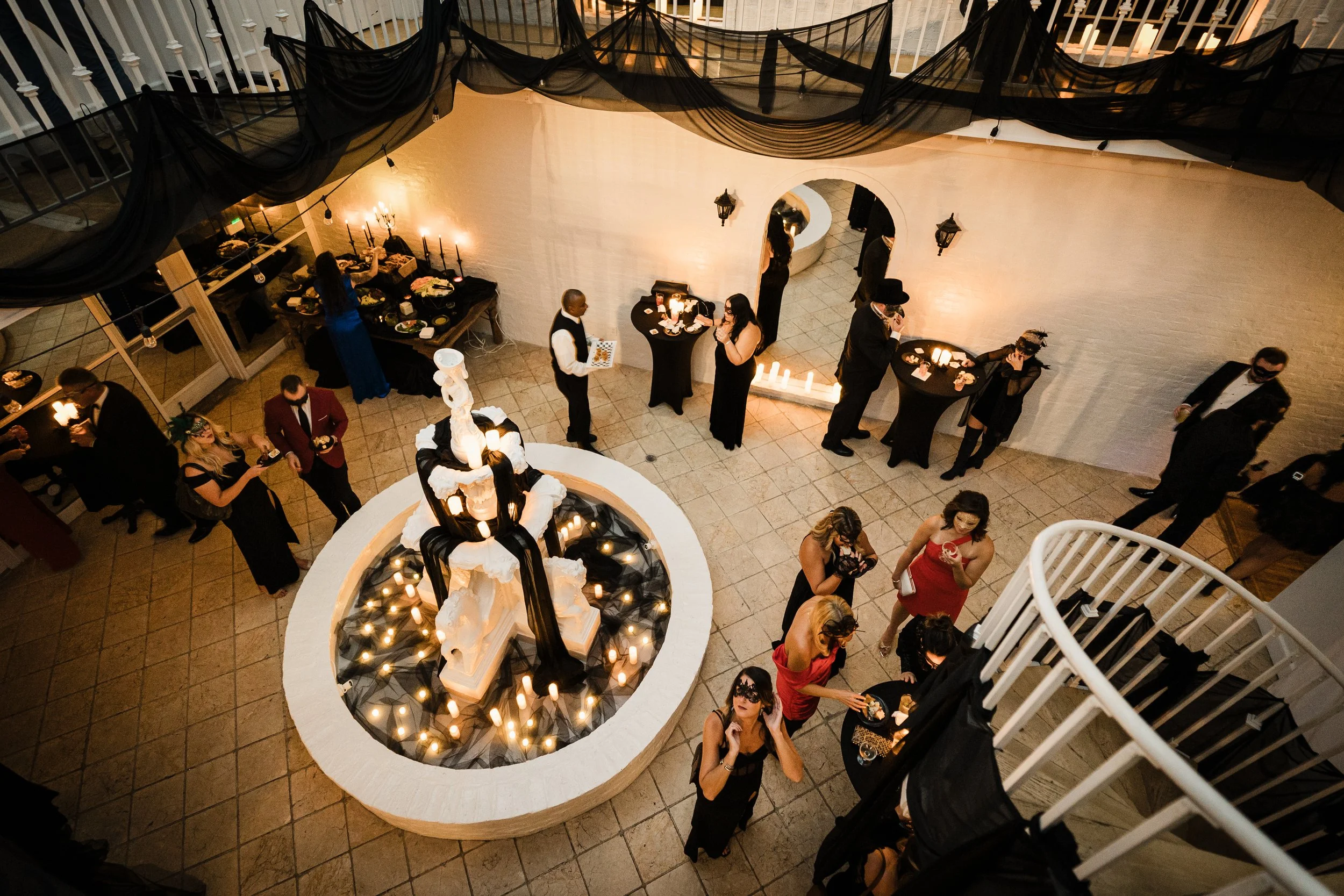 Indoor party scene viewed from above, featuring guests in formal attire socializing around cocktail tables and a white spiral staircase, with black drapery and lit candles decor.